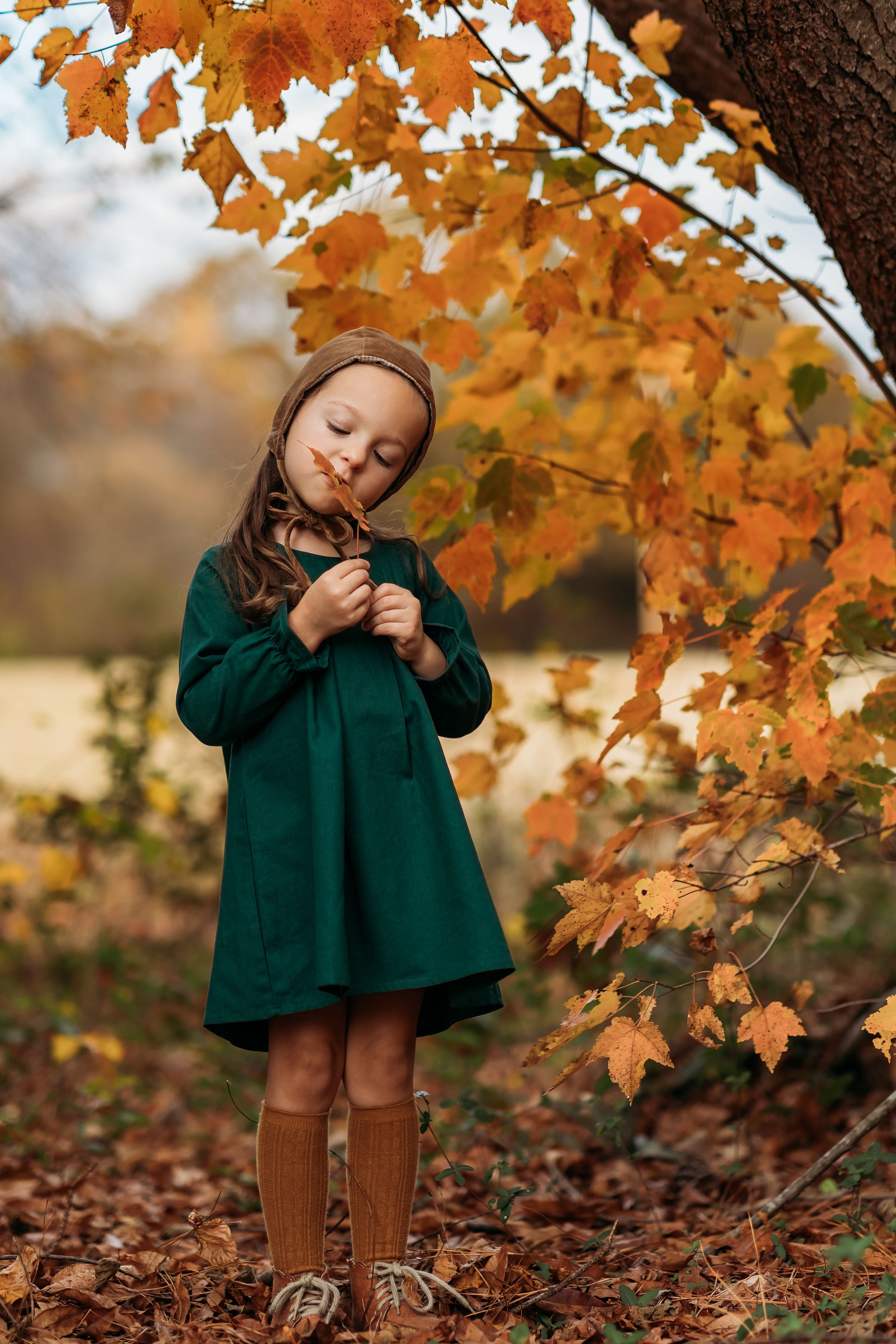 A 4-year-old girl standing in a fall setting, wearing the evergreen linen dress while holding a brown leaf, capturing the seasonal charm and style of the outfit.