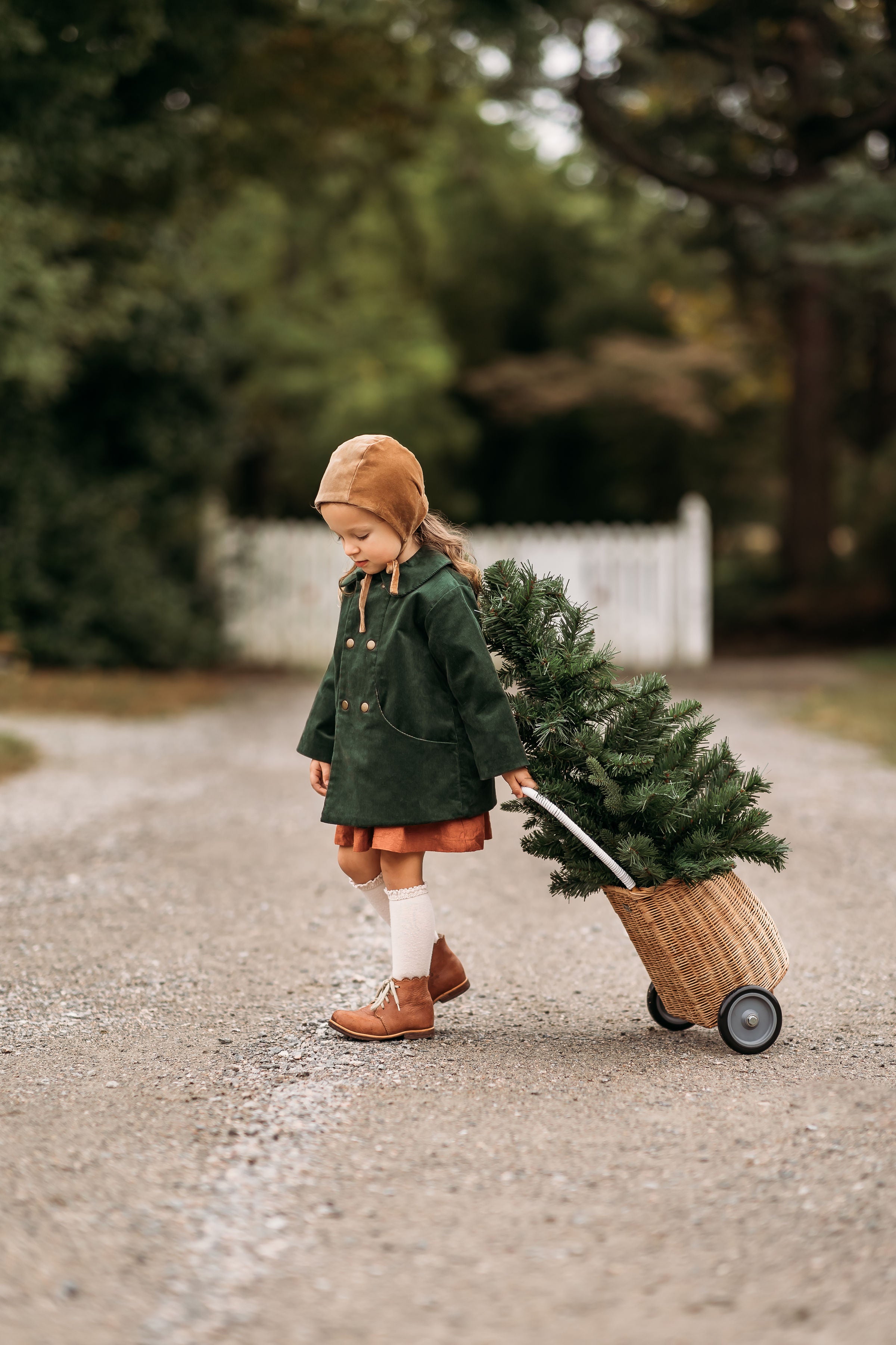 Side view of a girl in the Gold Velvet Bonnet and Evergreen Corduroy Coat, walking away with a Christmas tree, illustrating the bonnet’s elegant style and practicality for special celebrations.