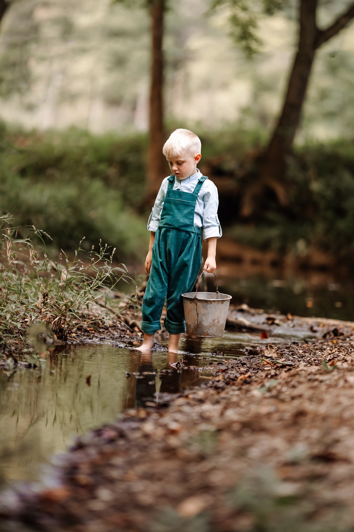 Child in green overalls holding a bucket by a stream in a forest