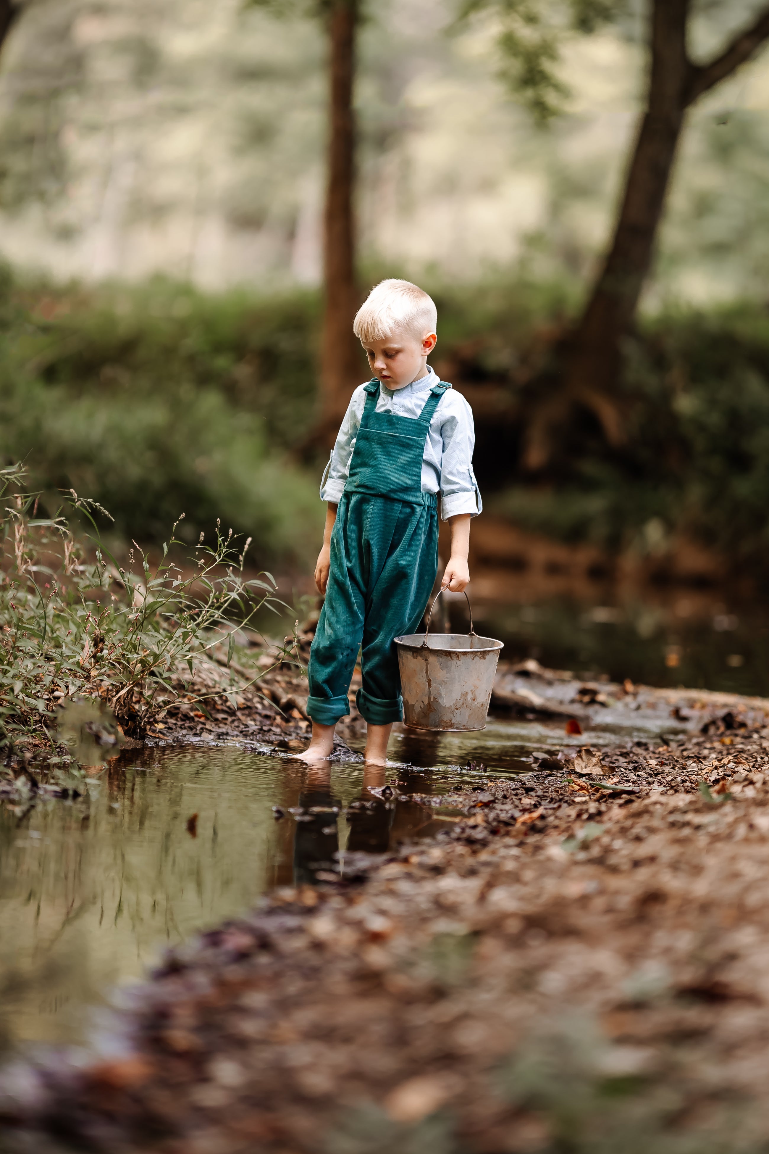 Child in green overalls holding a bucket by a stream in a forest