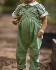 A toddler standing outdoors on a stone path wearing sage green linen-style overalls over a green and white gingham shirt with a white collar.