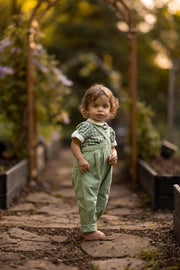 A toddler standing barefoot on a stone path in a garden, wearing sage green overall-style pants and a green-and-white checkered short-sleeve top.