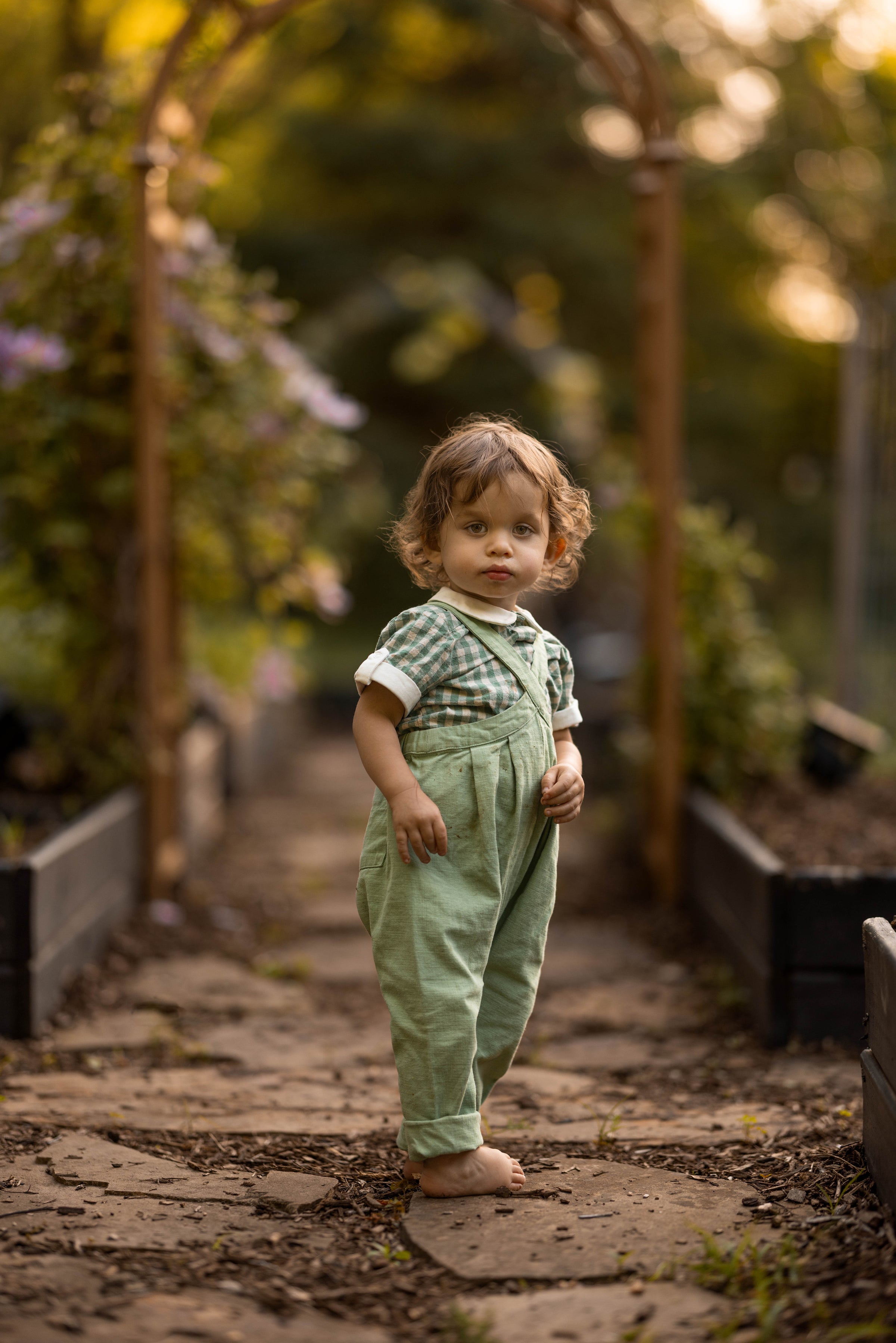 A toddler standing barefoot on a stone path in a garden, wearing sage green overall-style pants and a green-and-white checkered short-sleeve top.
