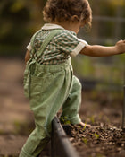 A toddler seen from behind wearing sage green linen overall pants and a green-and-white gingham shirt, barefoot in a garden.