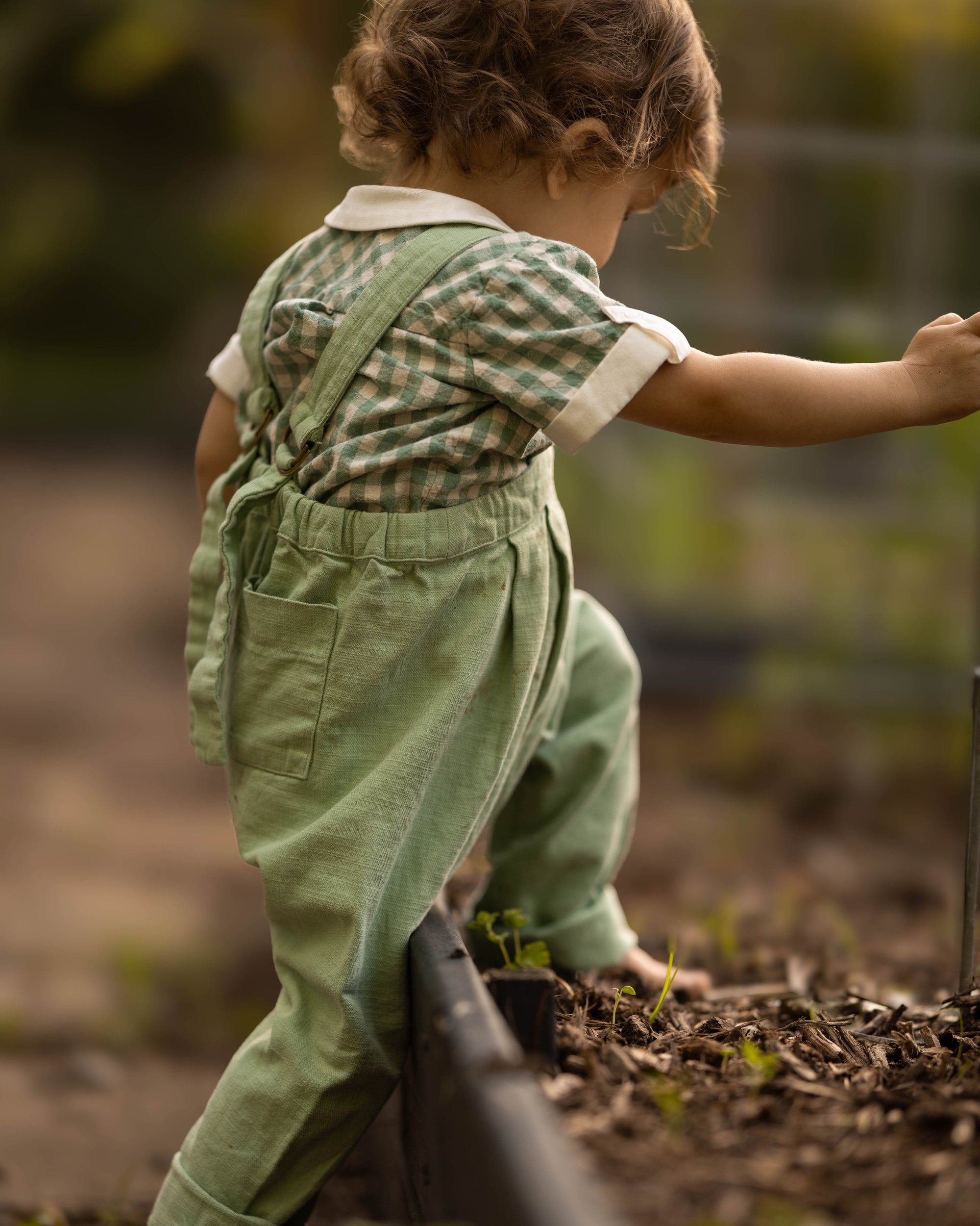 A toddler seen from behind wearing sage green linen overall pants and a green-and-white gingham shirt, barefoot in a garden.