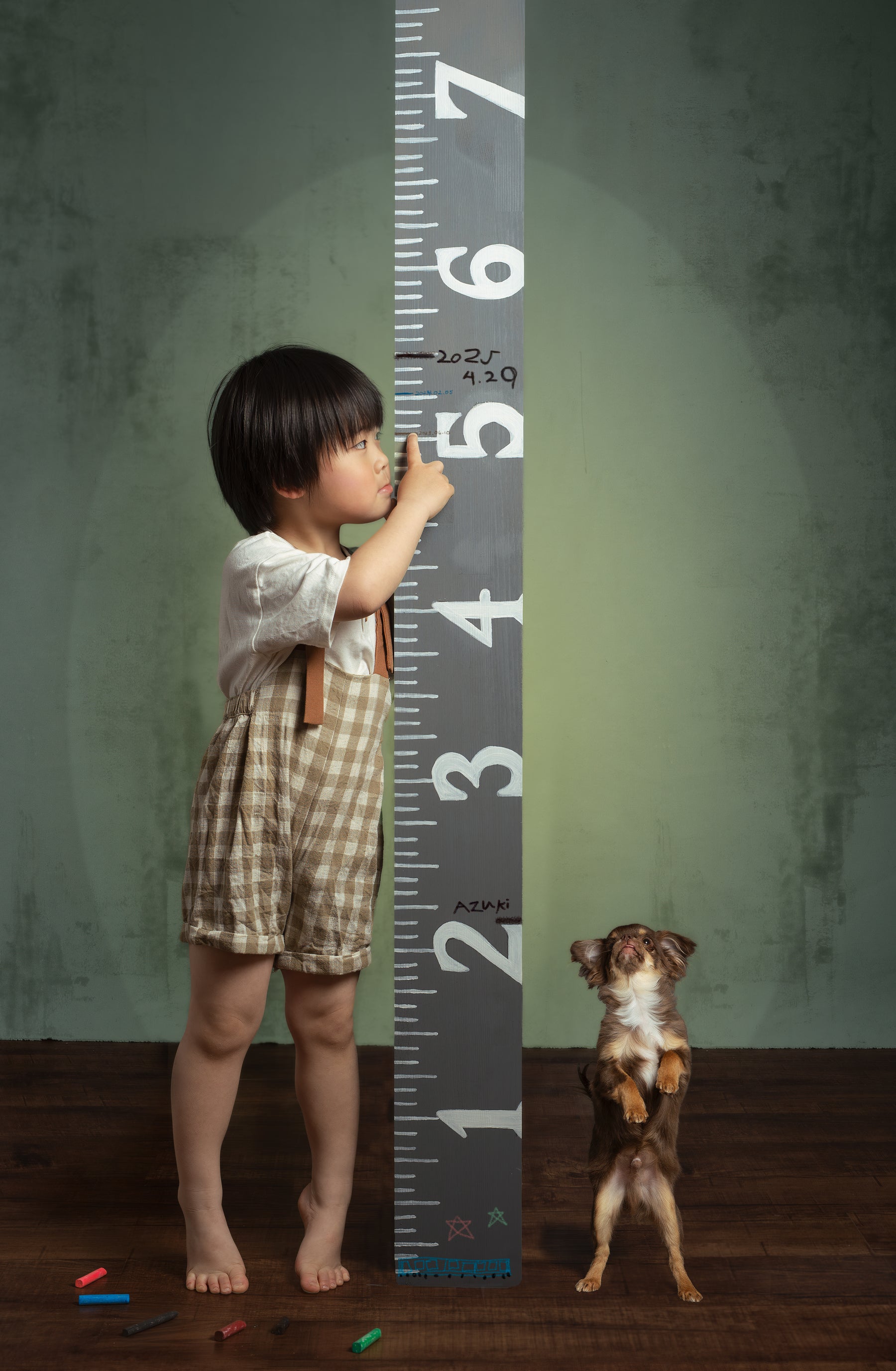 Child standing next to a large ruler with a dog at the bottom, against a textured wall.
