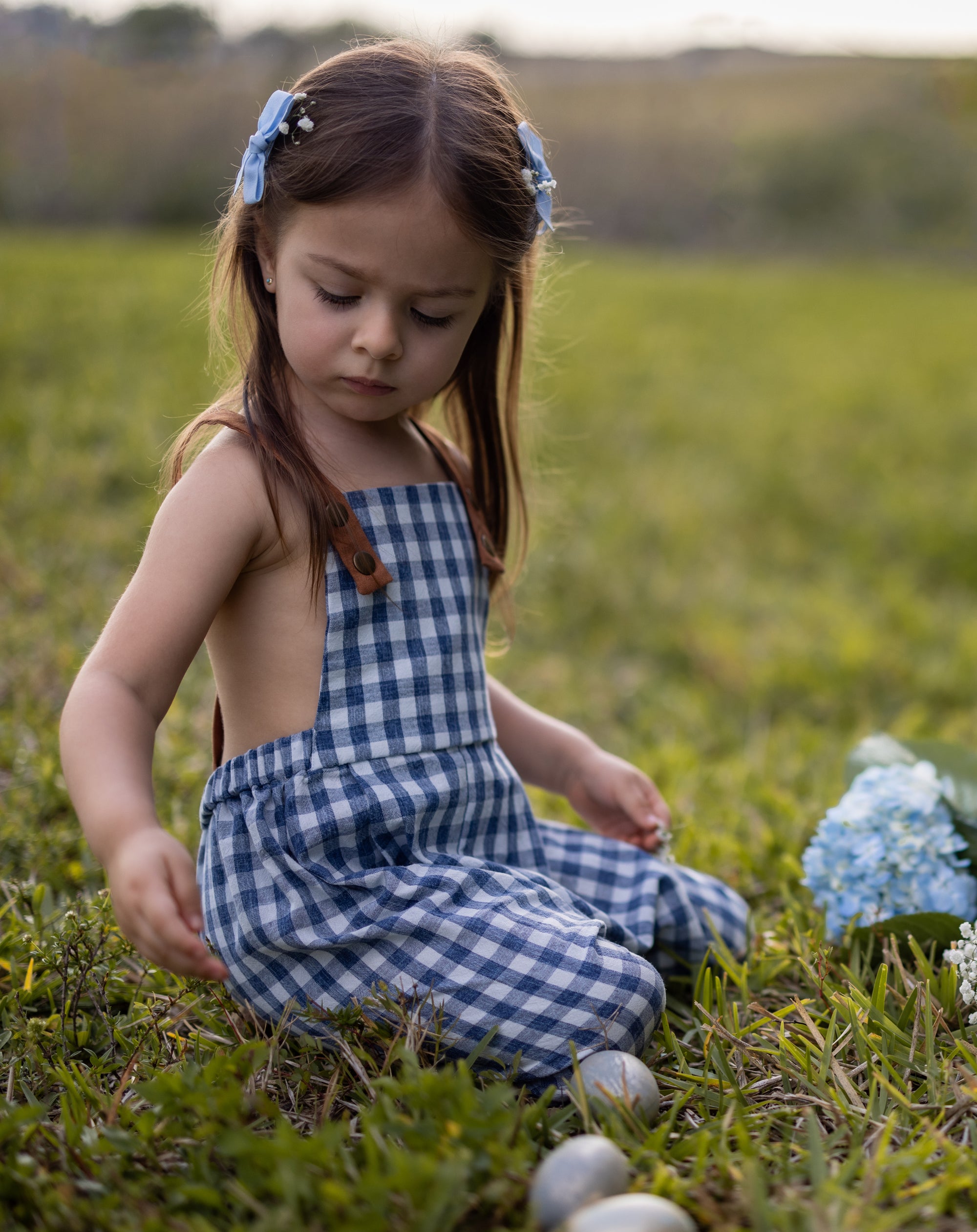A little girl sitting down and playing while wearing Blue Gingham Linen Long Overalls, enjoying her Easter festivities. The gender-neutral design showcases her playful spirit, while the breathable fabric ensures comfort during her activities. Stylish pleats enhance the overall look, and the durable leather-looking straps along with an adjustable fit make these overalls perfect for active little ones, all by a brand dedicated to sustainable fashion.