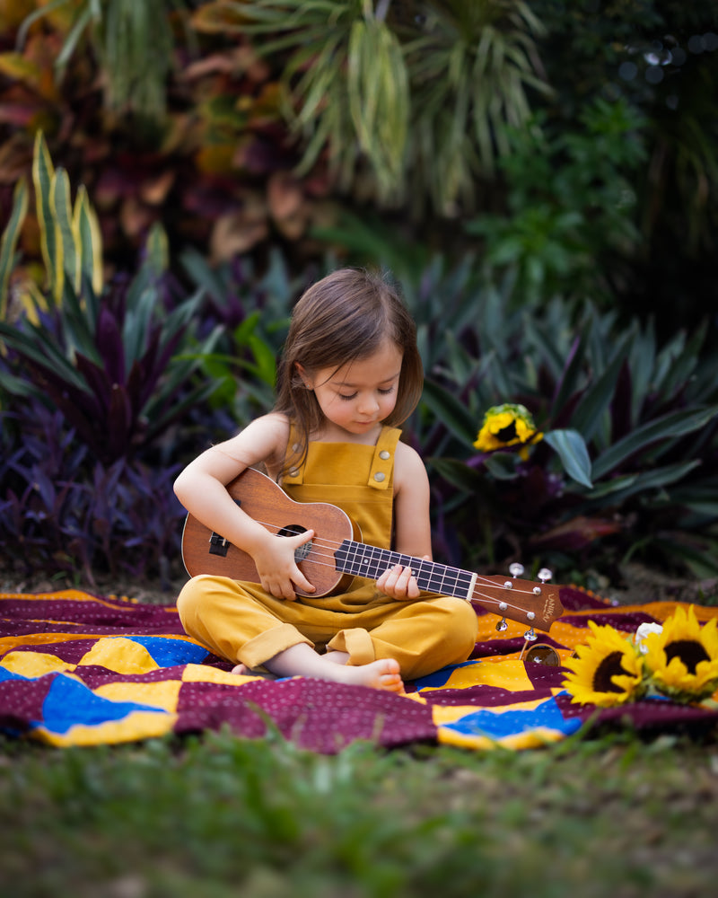 A 4-year-old girl playing ukulele freely in ginger long overalls, emphasizing the comfort and durability of the handmade 100% cotton fabric.