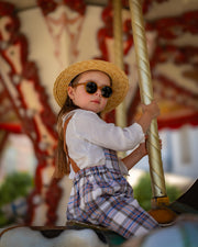 Child on a carousel holding onto a pole, wearing a straw hat and sunglasses and plaid linen overalls.