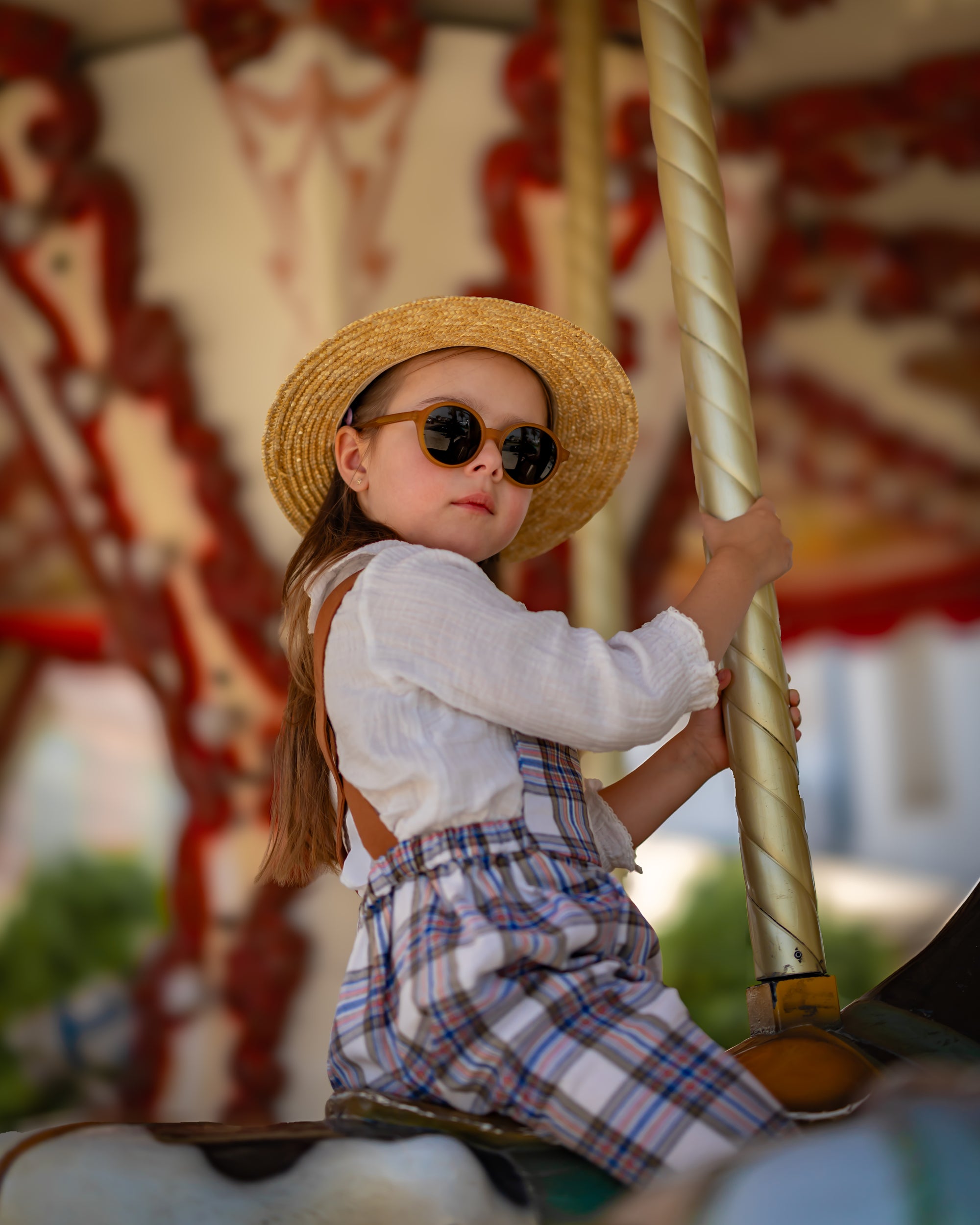 Child on a carousel holding onto a pole, wearing a straw hat and sunglasses and plaid linen overalls.