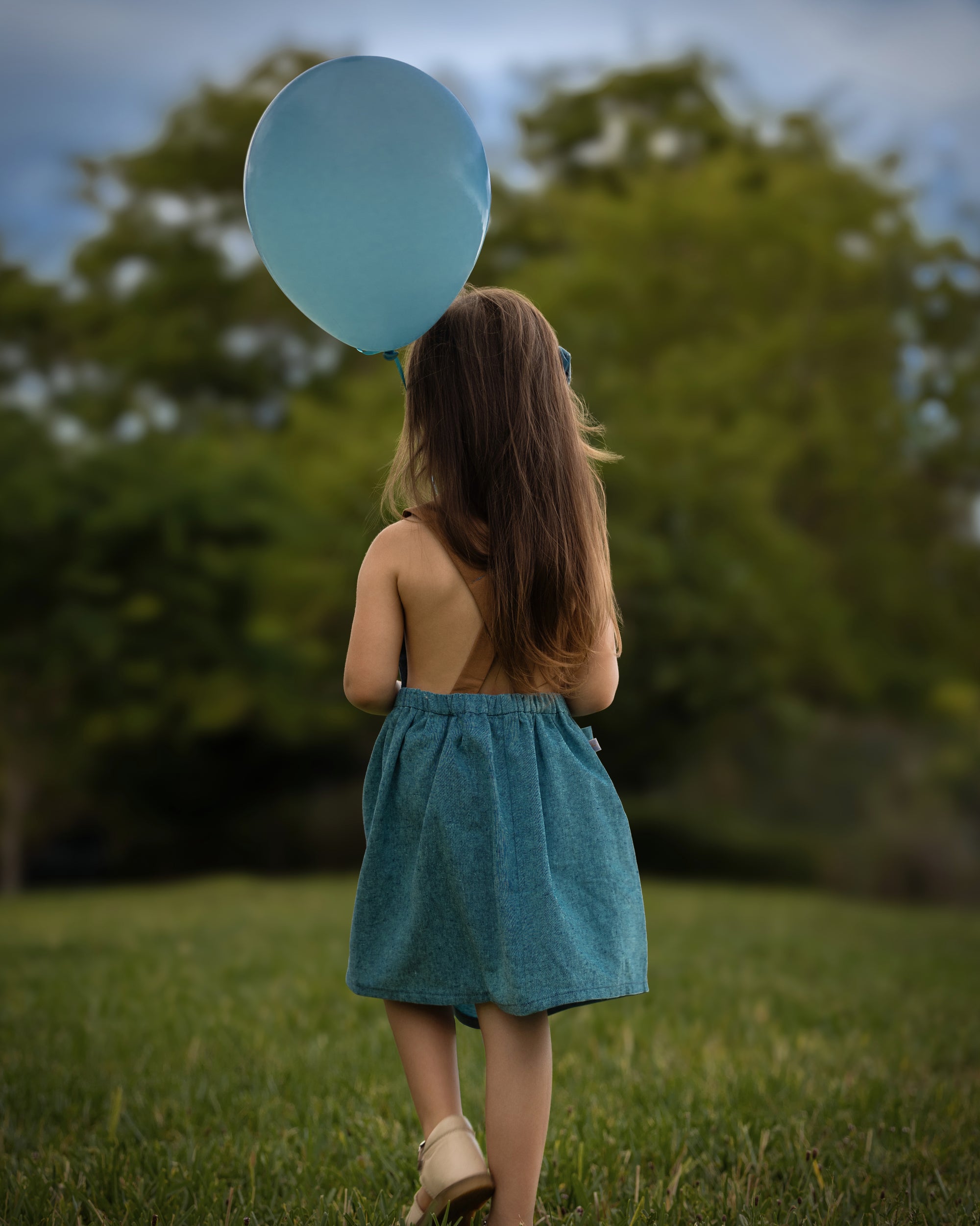 A little girl model walking away in the Blue Jay pinafore, showcasing the back in a summer setting - Handmade from repurposed linen in our NYC studio, this classic design features durable leather-looking straps and adjustable lengths for a perfect fit. The generous pocket is great for storing little treasures, making it a stylish choice for summer adventures.