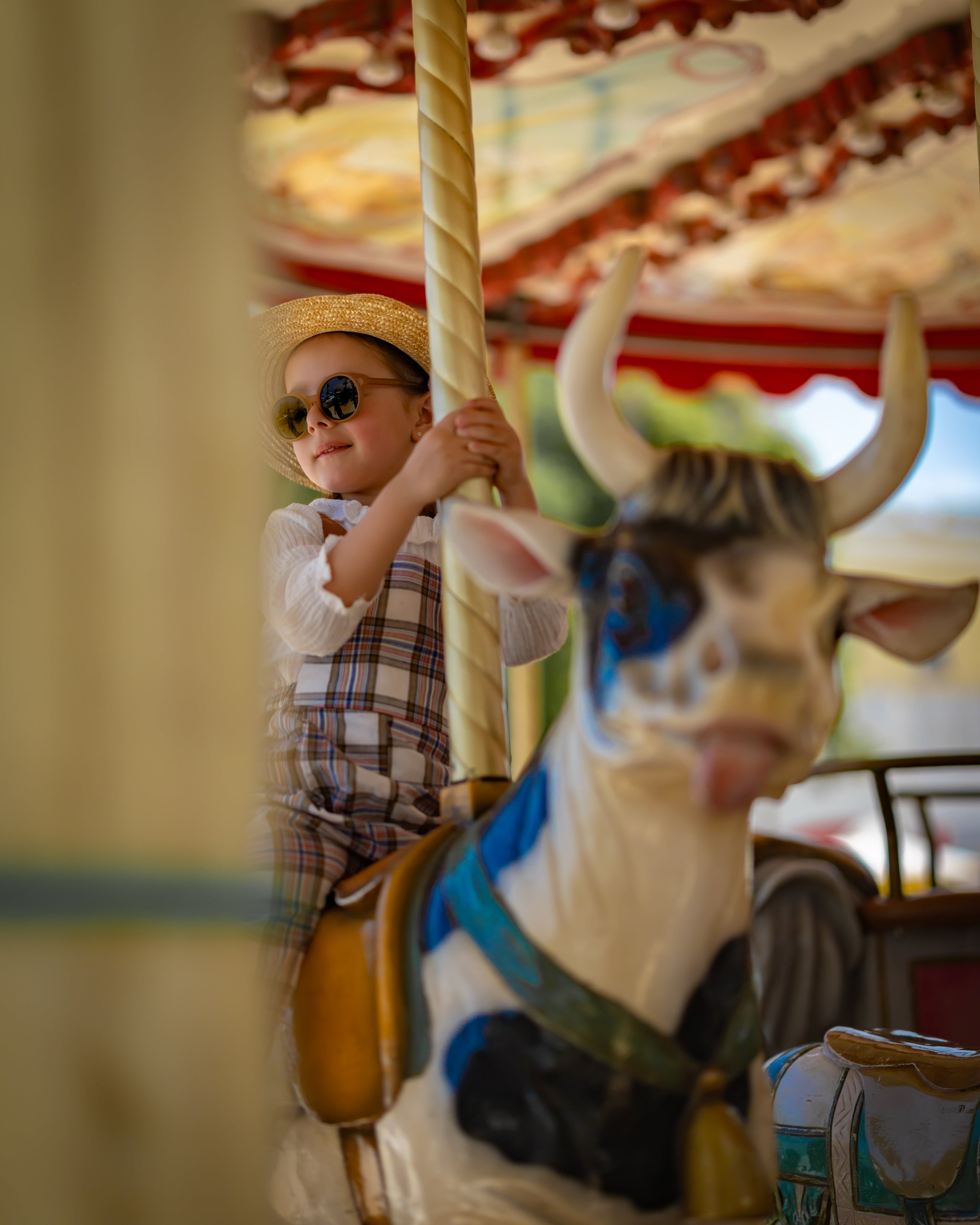Child on a carousel with a cow figure, wearing sunglasses and a hat and plaid linen overalls.