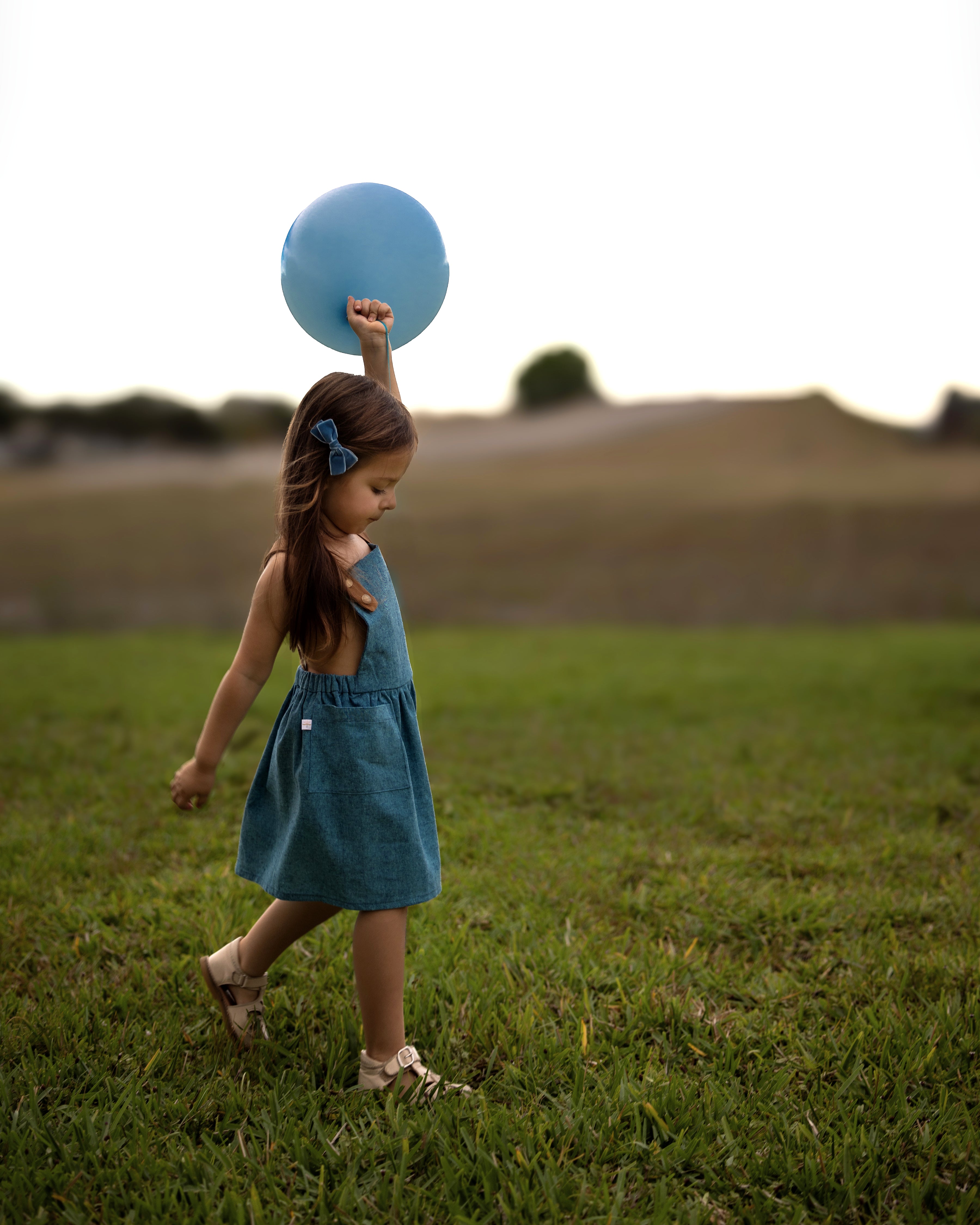 Side view of a little girl model wearing the Blue Jay pinafore while holding a blue balloon in the summer - This classic and timeless design is handmade from repurposed linen in our NYC studio. The pinafore is designed to be worn on its own, allowing your little girl to stay stylish and cool. The durable leather-looking straps add visual appeal, while the adjustable straps in two lengths ensure a perfect fit. A generous pocket is perfect for storing little treasures, making it an ideal choice for summer fun