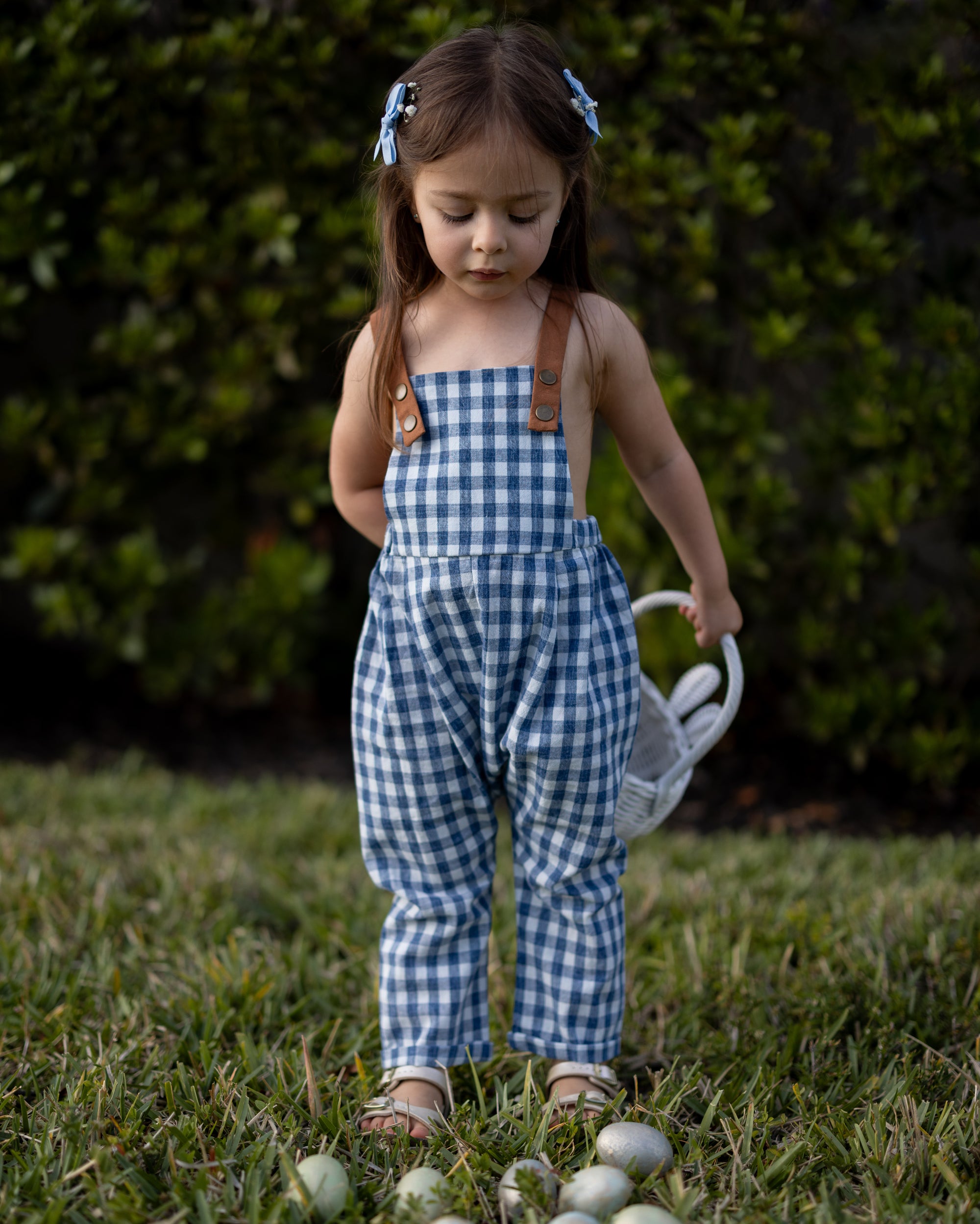 A little girl joyfully wearing Blue Gingham Linen Long Overalls, showcasing the playful and gender-neutral design perfect for Easter celebrations. The breathable fabric keeps her comfortable, while stylish pleats add a charming touch. The durable leather-looking straps and adjustable fit ensure she looks adorable and feels great during her festive activities.