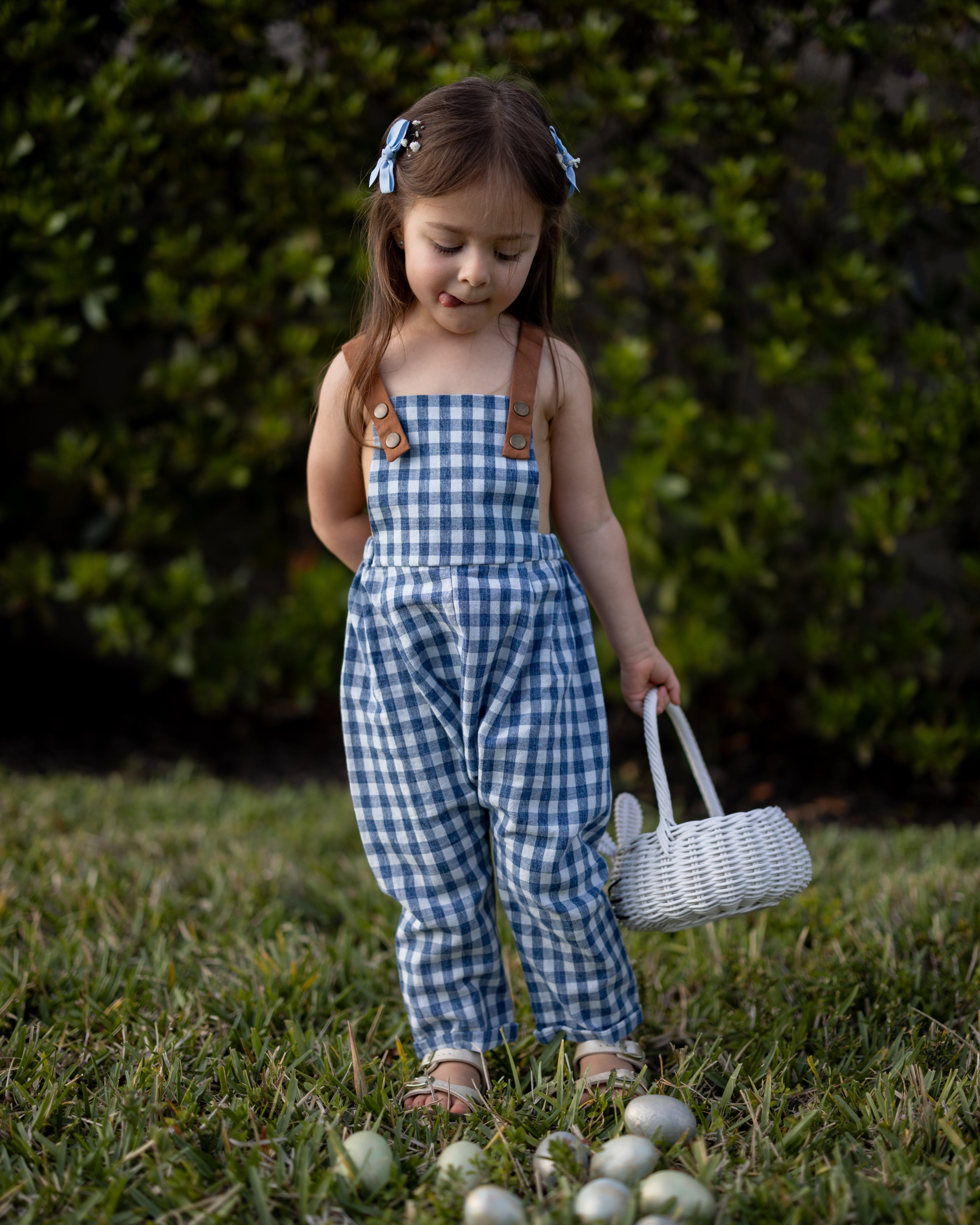 A little girl playfully sticking her tongue out while wearing Blue Gingham Linen Long Overalls, capturing a joyful moment during Easter. The gender-neutral design and breathable fabric allow for carefree movement, while the stylish pleats add a touch of elegance. Durable leather-looking straps and an adjustable fit make these overalls both fun and practical, reflecting the brand's commitment to sustainable fashion for children.