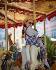 Child on a carousel with colorful background wearing a hat and plaid linen overalls.