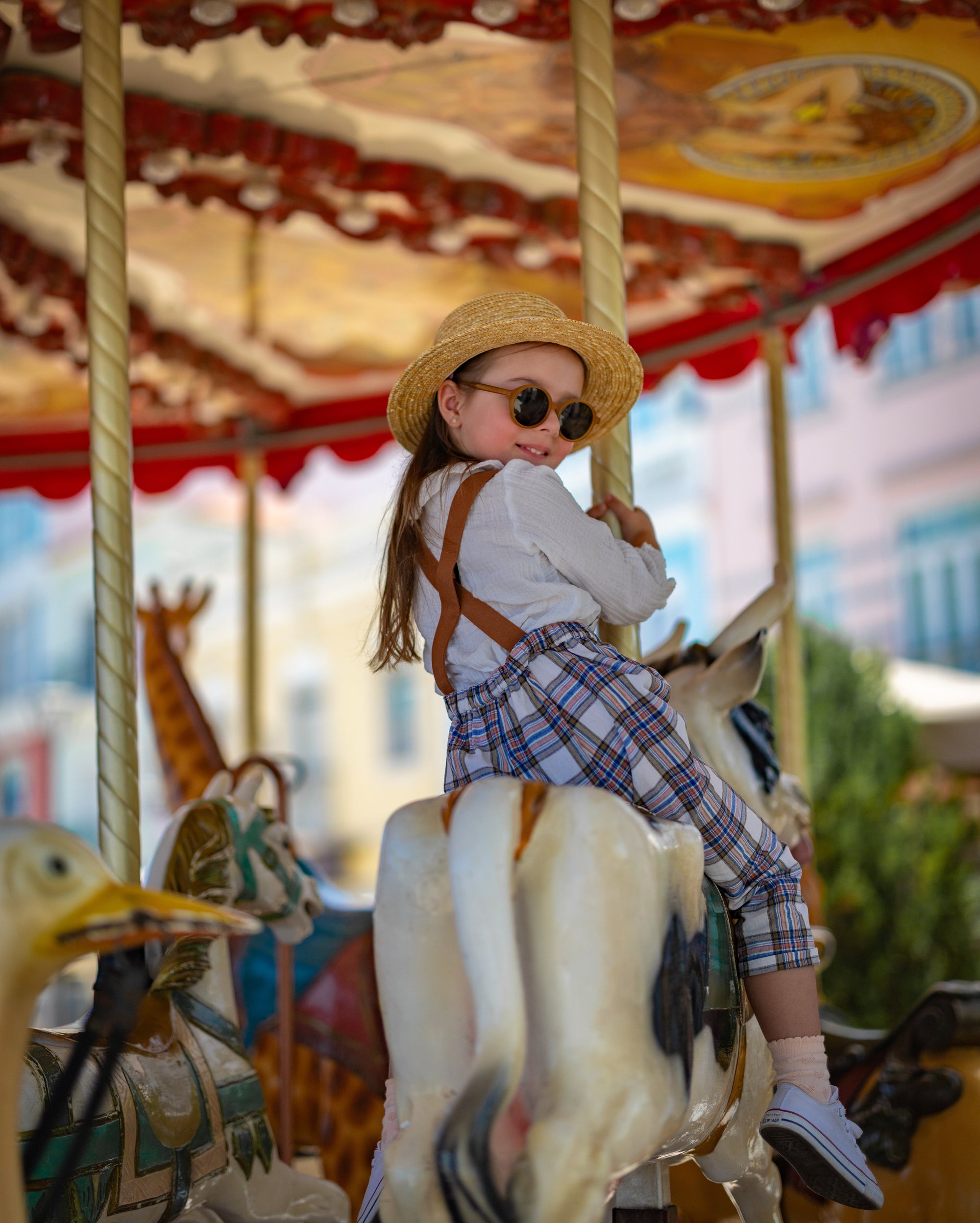 Child on a carousel with colorful background wearing a hat and plaid linen overalls.