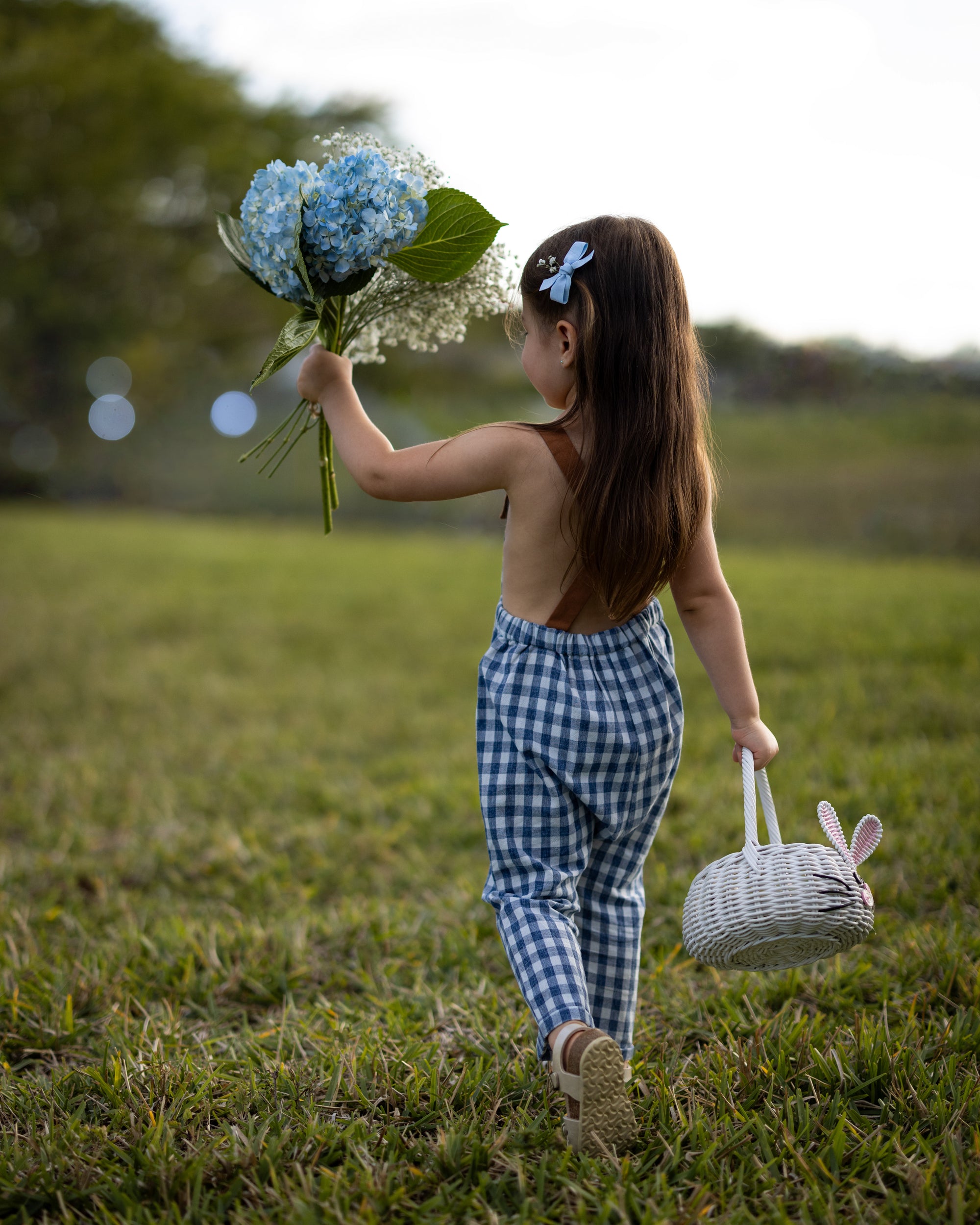 Blue Gingham Linen Long Overalls on a little girl from the back during Easter, showcasing the gender-neutral design perfect for passing down, breathable fabric for all-day comfort, stylish pleats for elegance, durable leather-looking straps, and adjustable straps for a perfect fit. Made by a sustainable brand committed to eco-friendly practices, promoting responsible fashion for children.