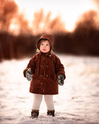 Little girl in the snow in a rust coat and matching hat with blurry background