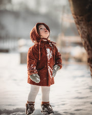 Little girl in the snow in a rust coat and matching hat looking up