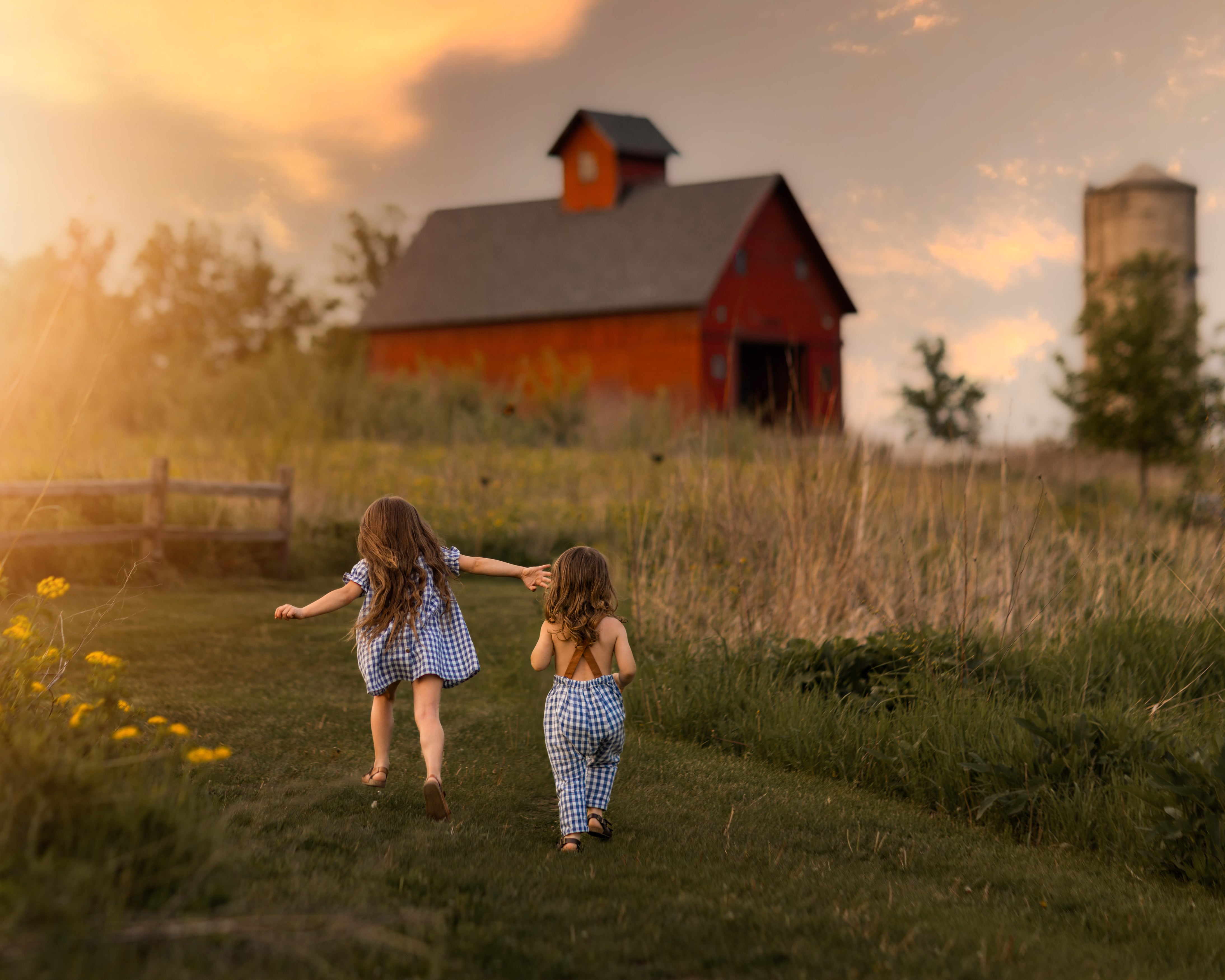 The little girl and her brother are seen from the back, running side by side towards the sunset. The girl wears her Blue Gingham linen dress, which sways with her movement, while her brother sports matching blue gingham overalls. The warm, golden light of the setting sun illuminates their figures, capturing the essence of carefree childhood as they enjoy the moment together in this enchanting outdoor setting.