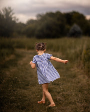 A little girl stands with her back to the camera, showcasing the Blue Gingham linen dress. The dress features a pin-tucked front and a yoke with two pleats at the back, emphasizing its charming design. The delicate ruffles at the short sleeve hem add a sweet touch, while the A-line shape allows for comfortable movement. Her hair is styled up, and she enjoys the sunny day in a picturesque field, highlighting the dress's practicality and style.