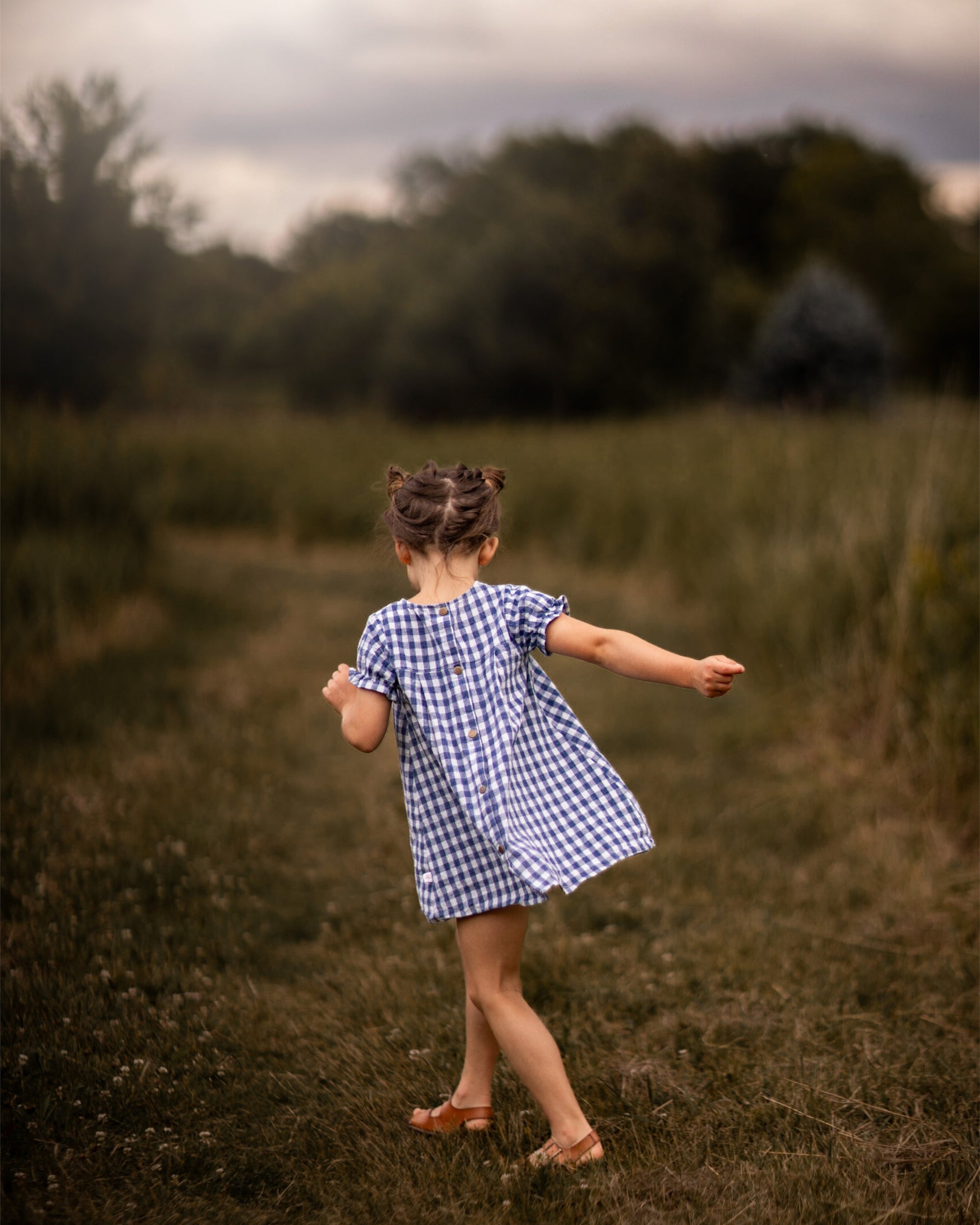 A little girl stands with her back to the camera, showcasing the Blue Gingham linen dress. The dress features a pin-tucked front and a yoke with two pleats at the back, emphasizing its charming design. The delicate ruffles at the short sleeve hem add a sweet touch, while the A-line shape allows for comfortable movement. Her hair is styled up, and she enjoys the sunny day in a picturesque field, highlighting the dress's practicality and style.