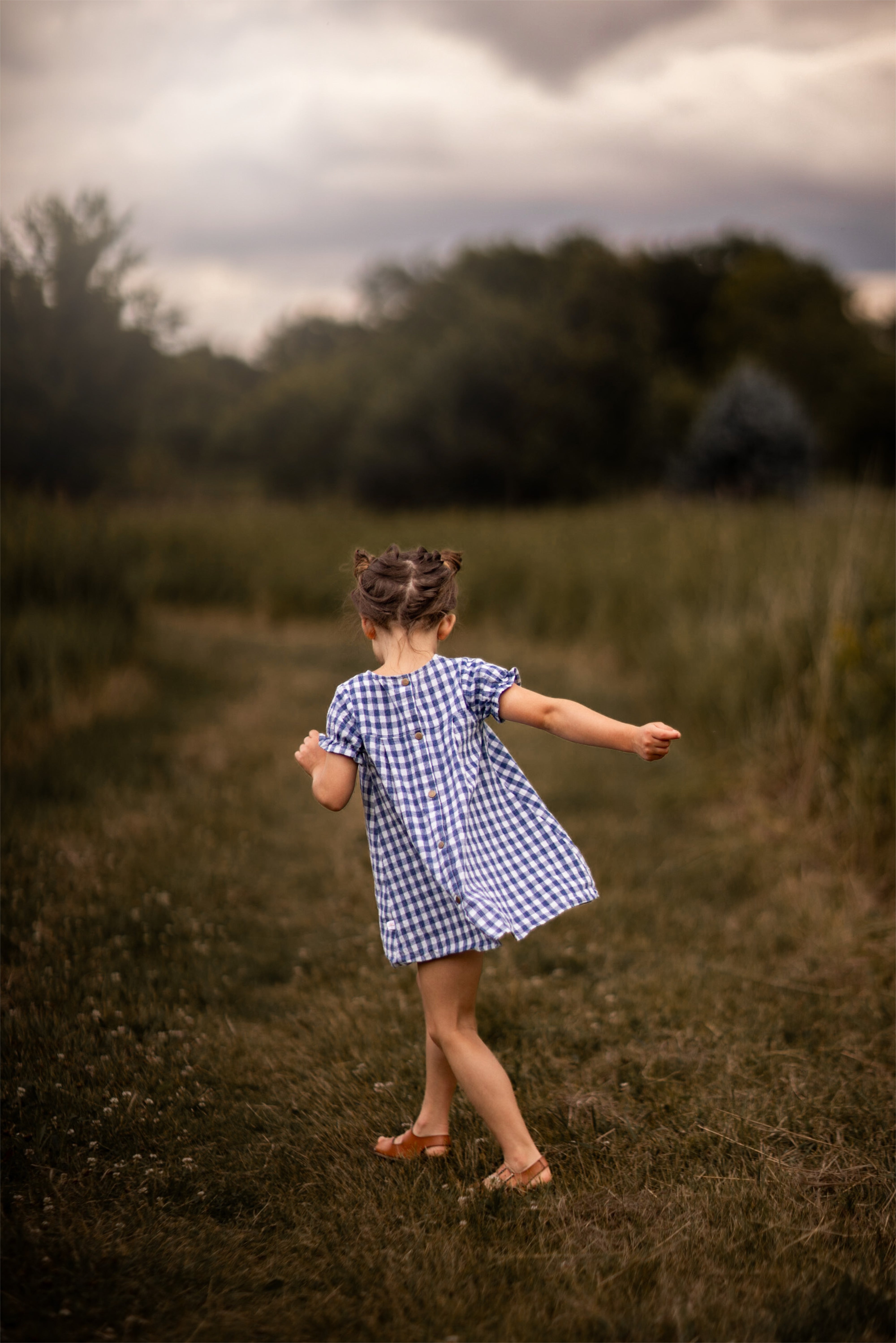 A little girl stands with her back to the camera, showcasing the Blue Gingham linen dress. The dress features a pin-tucked front and a yoke with two pleats at the back, emphasizing its charming design. The delicate ruffles at the short sleeve hem add a sweet touch, while the A-line shape allows for comfortable movement. Her hair is styled up, and she enjoys the sunny day in a picturesque field, highlighting the dress's practicality and style.