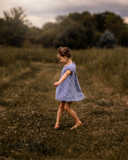 A little girl wearing a Blue Gingham linen dress dances joyfully in a field. Her hair is styled up, adding to her playful spirit. The dress features a pin-tucked front, a yoke with pleats at the back, and delicate ruffles at the short sleeve hem, creating a charming and stylish look. The A-line shape allows for easy movement, making it perfect for her carefree dancing in the warm summer breeze.