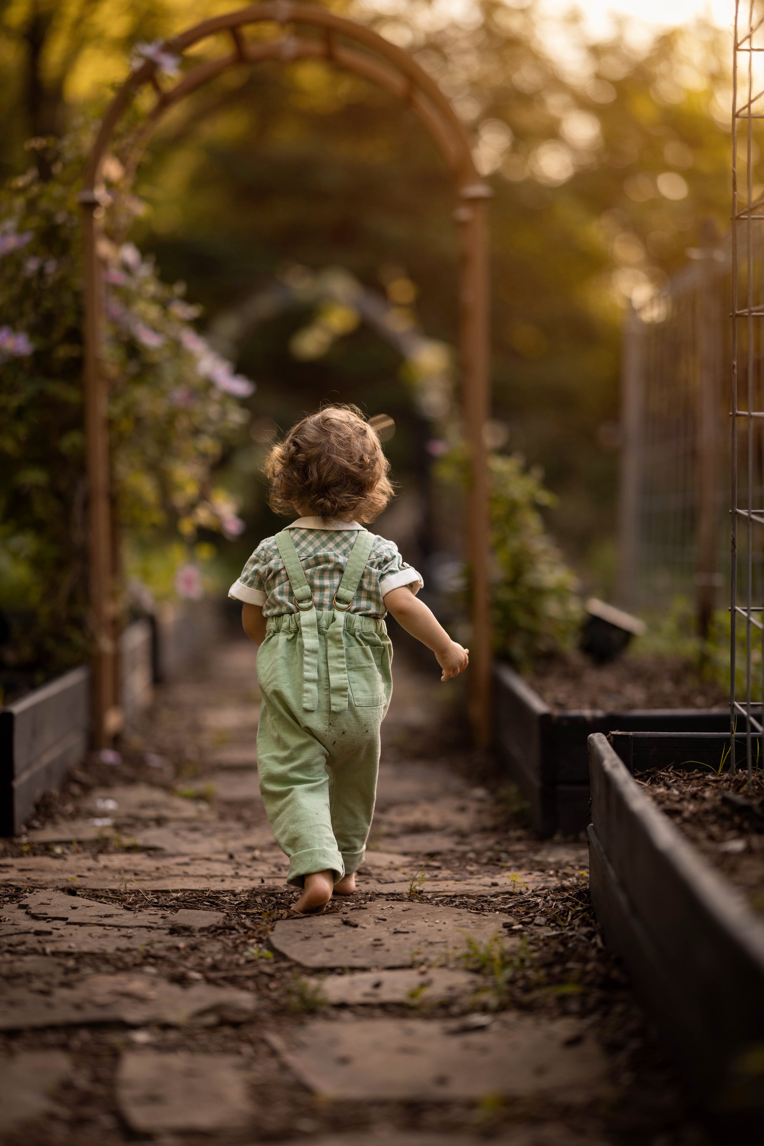 A toddler seen from behind walking barefoot down a garden path between raised garden beds, wearing a light green overall-style romper with a patterned short-sleeve top.