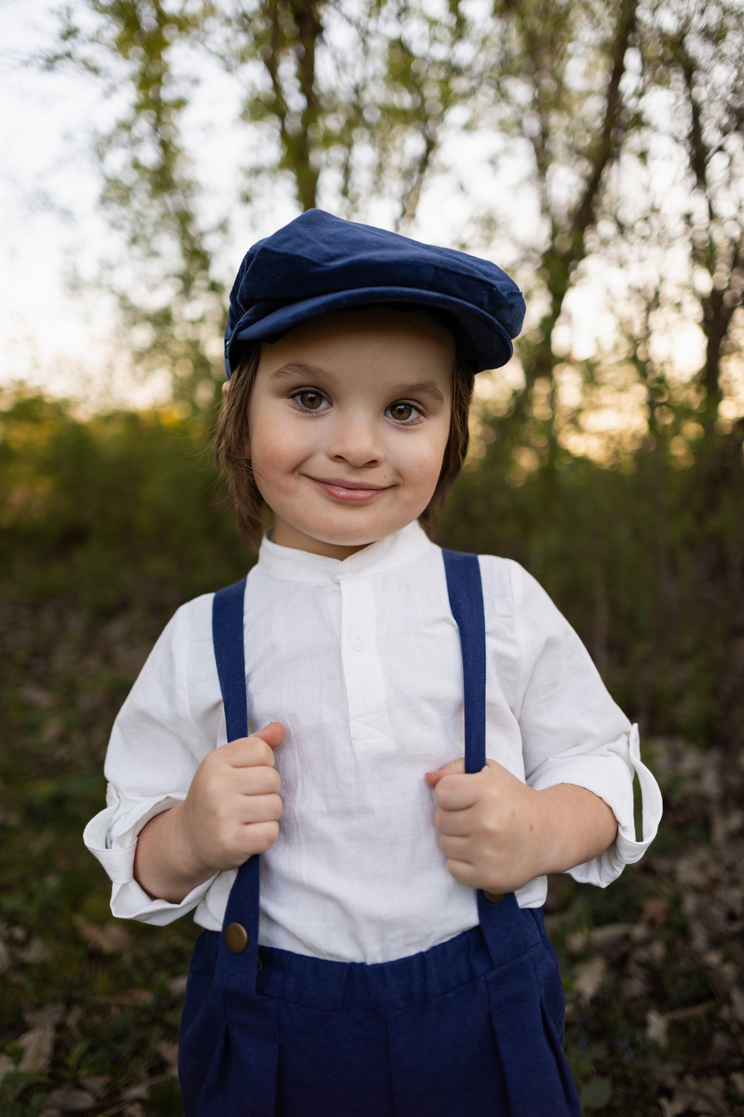 A close-up of boy, capturing his charming smirk and elegant features, highlighting the details of his outfit, including the navy linen suspender shorts and white Henley shirt, set against a soft background that enhances his stylish appearance.