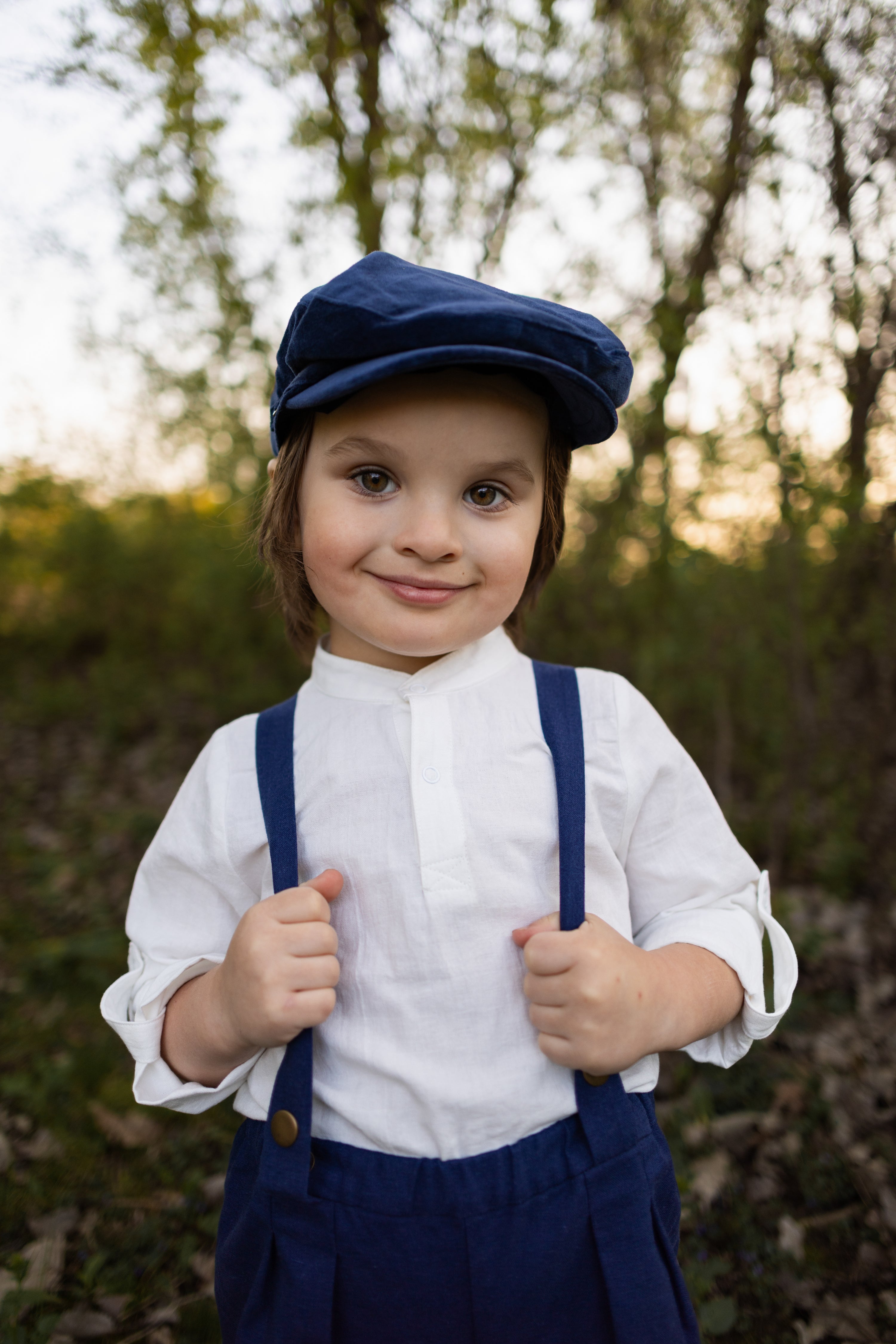 A close-up of the same model boy, capturing his charming smirk and elegant features, highlighting the details of his outfit, including the navy linen suspender shorts and white Henley shirt, set against a soft background that enhances his stylish appearance.