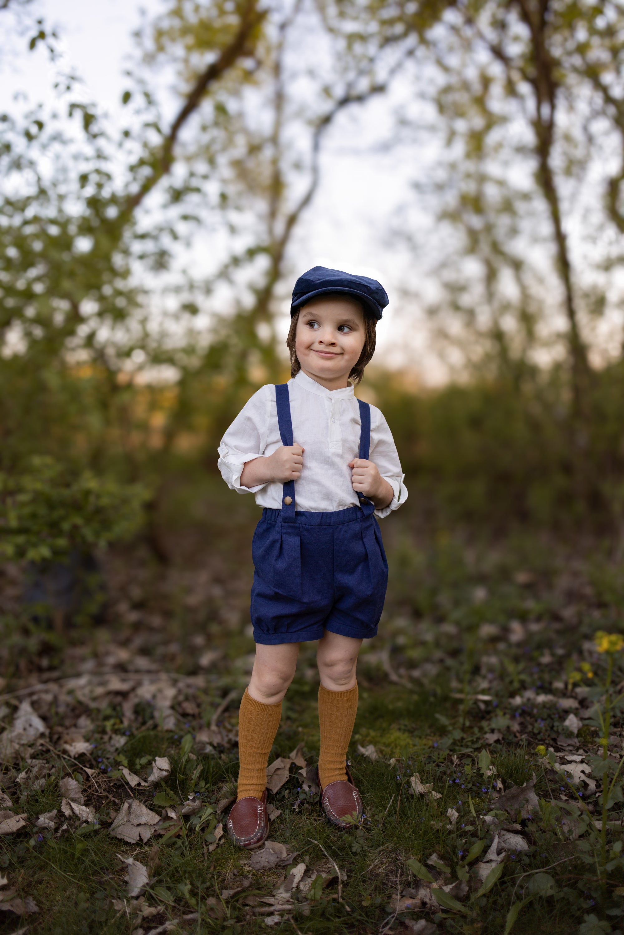 Child in vintage outfit with suspenders and cap standing in a forest