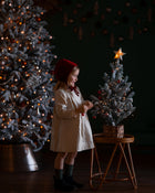 Child in a white dress and red hat standing next to a small Christmas tree with a star on top, in a dimly lit room.