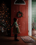 Girl standing in front of open door with snow flying in wearing a red velvet bonnet and a natural linen dress with a long green scarf