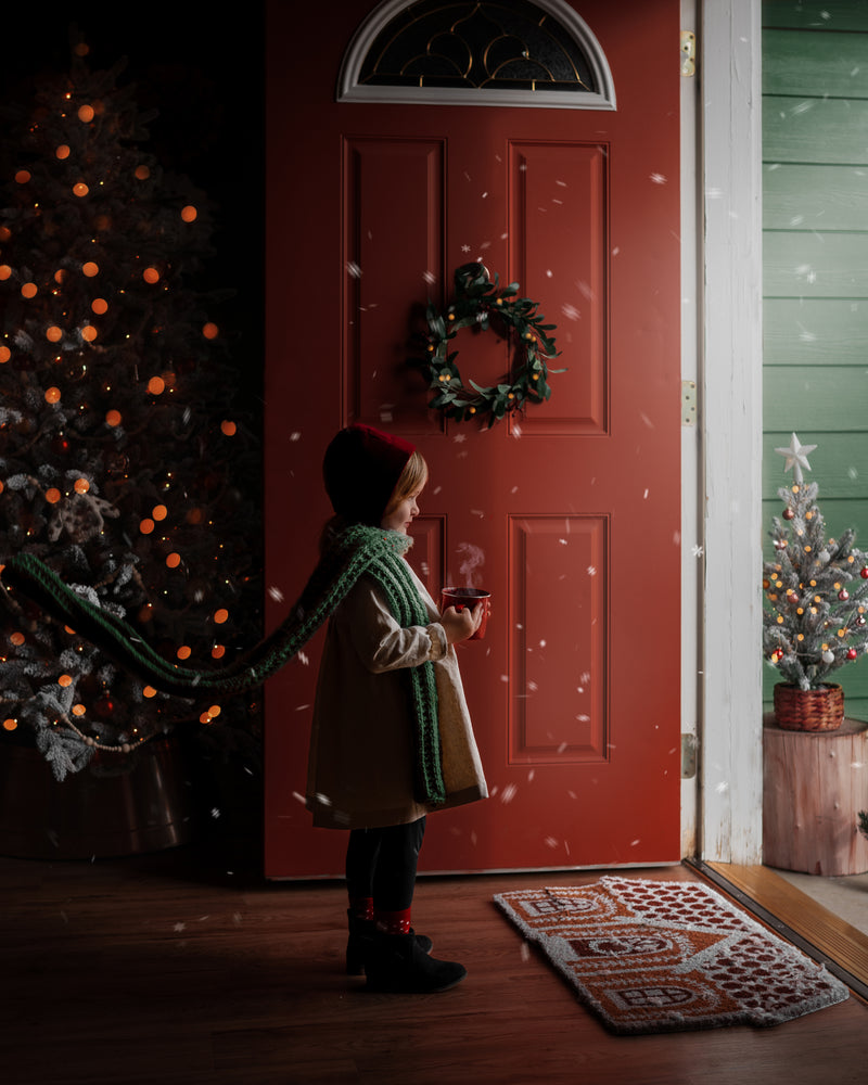 Girl standing in front of open door with snow flying in wearing a red velvet bonnet and a natural linen dress with a long green scarf