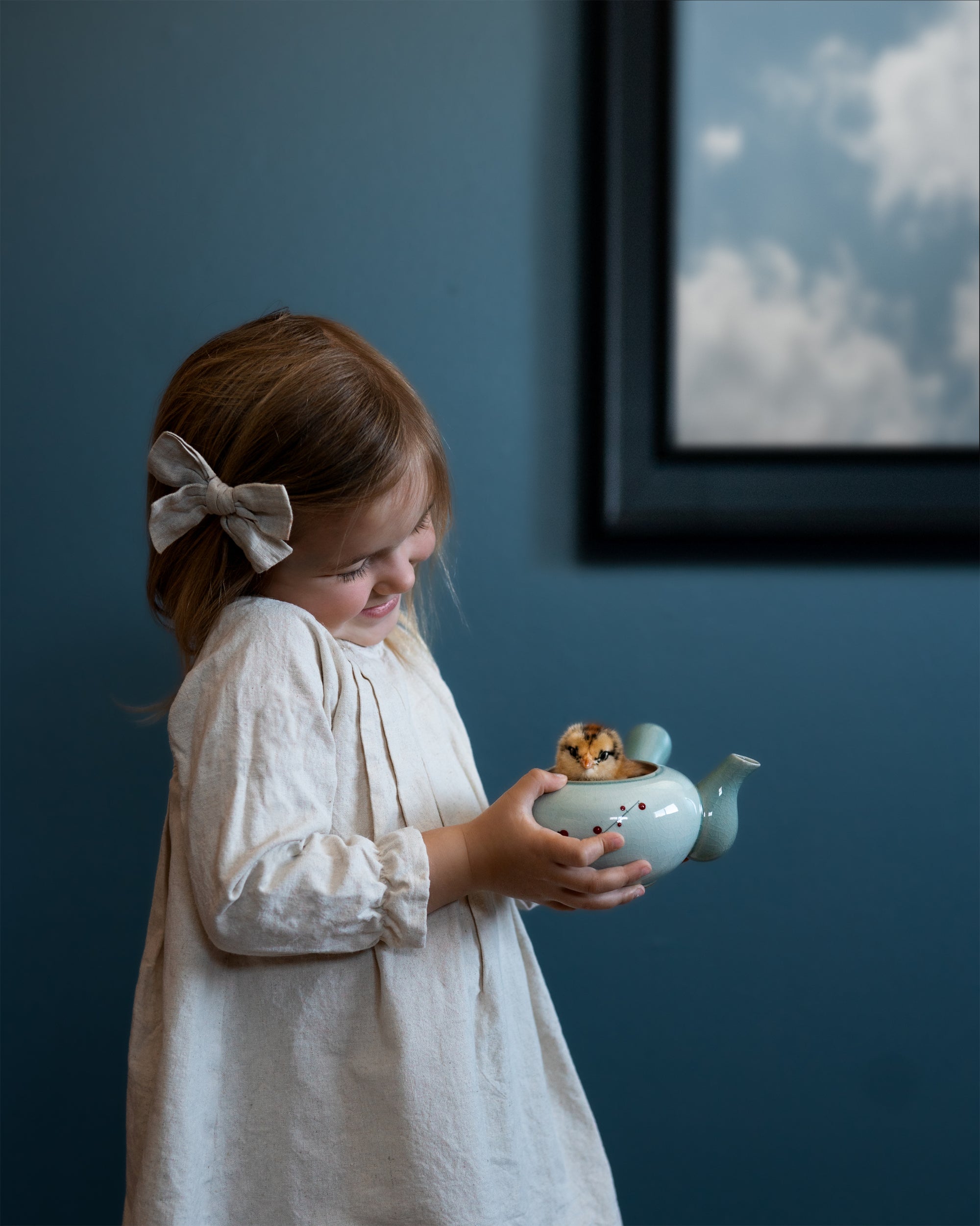 Young girl holding a small teapot with a bear figurine inside against a blue wall.