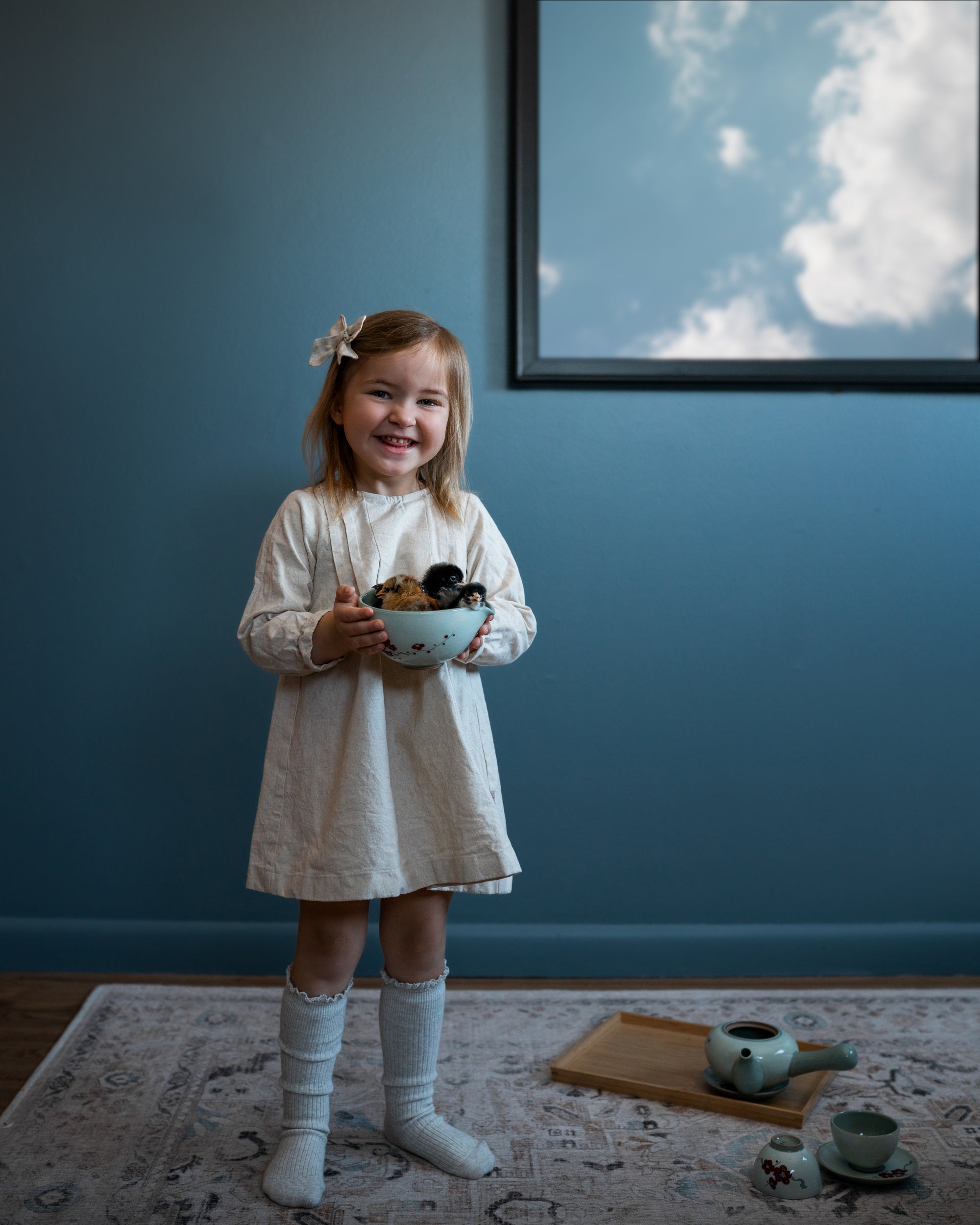 Young girl in a white dress holding a bowl against a blue wall with a framed picture.