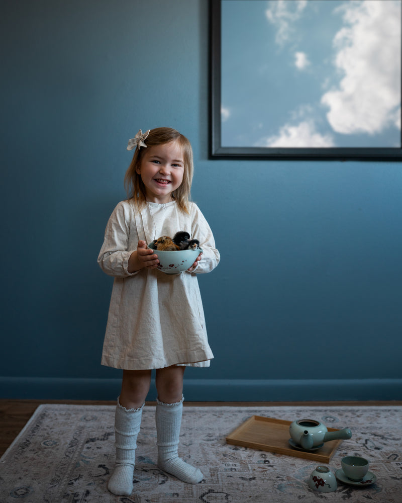 Young girl in a white dress holding a bowl against a blue wall with a framed picture.