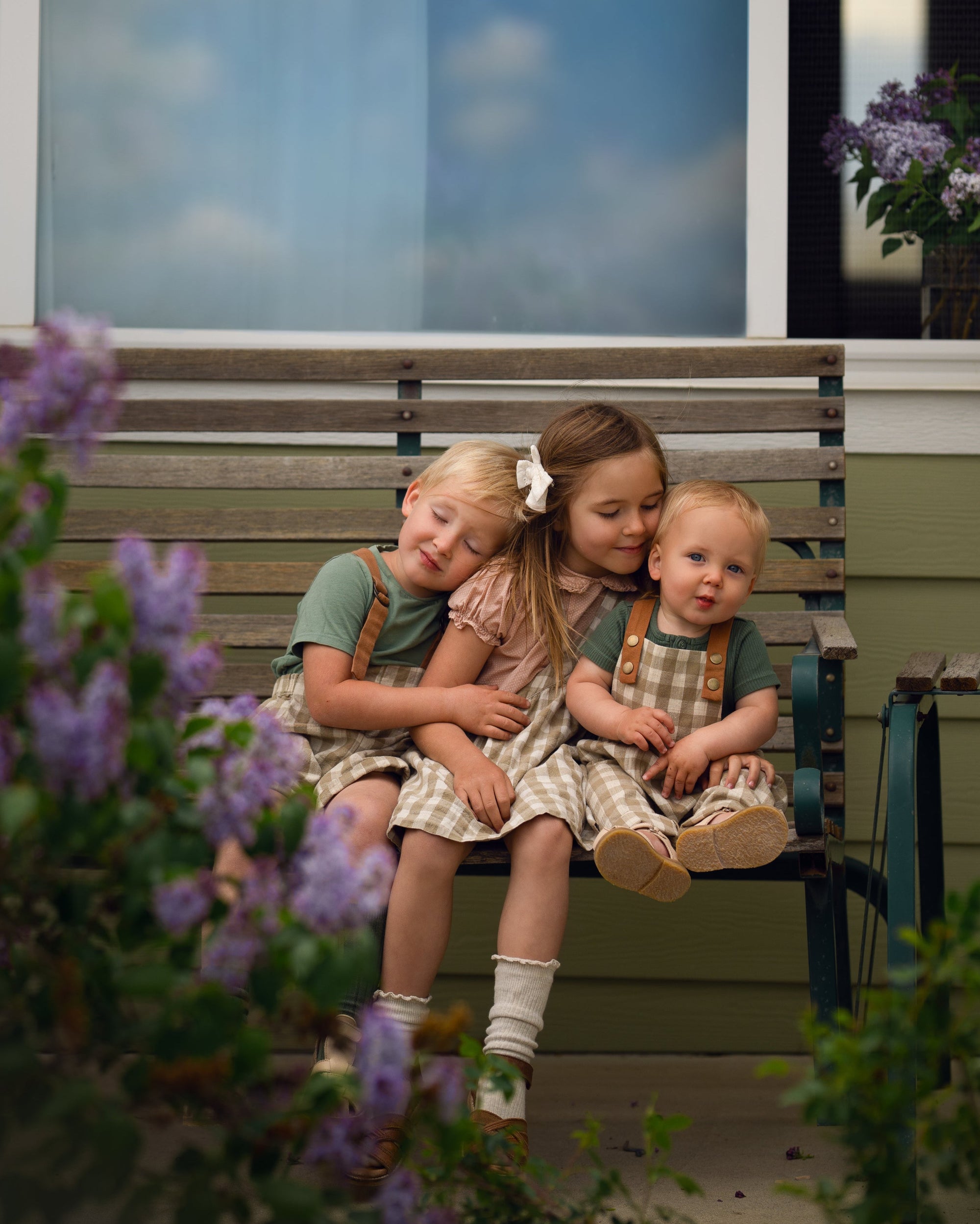 Three children sitting on a bench with flowers and a house in the background