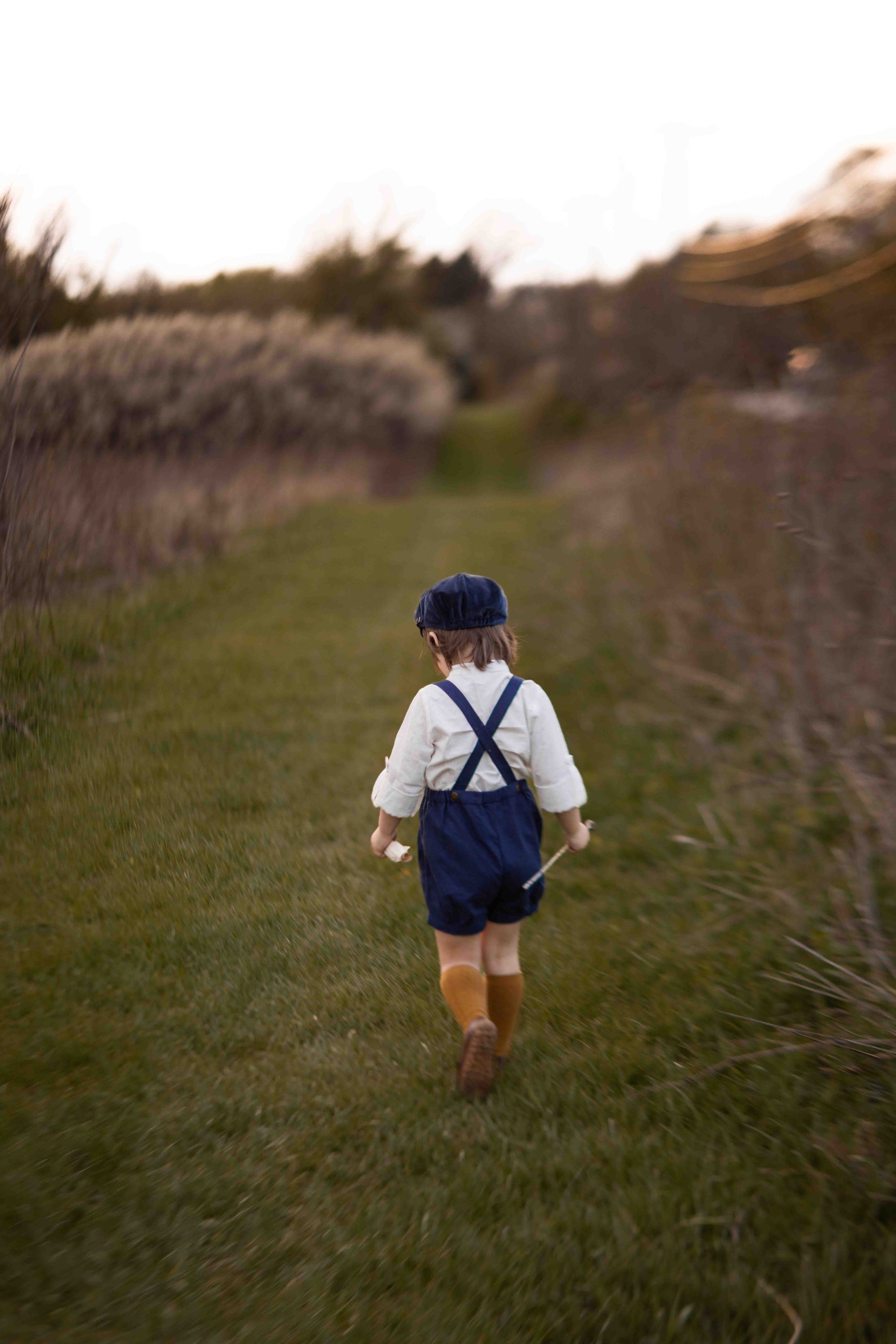 Child walking away in a field wearing a white shirt, blue overalls, and brown boots.
