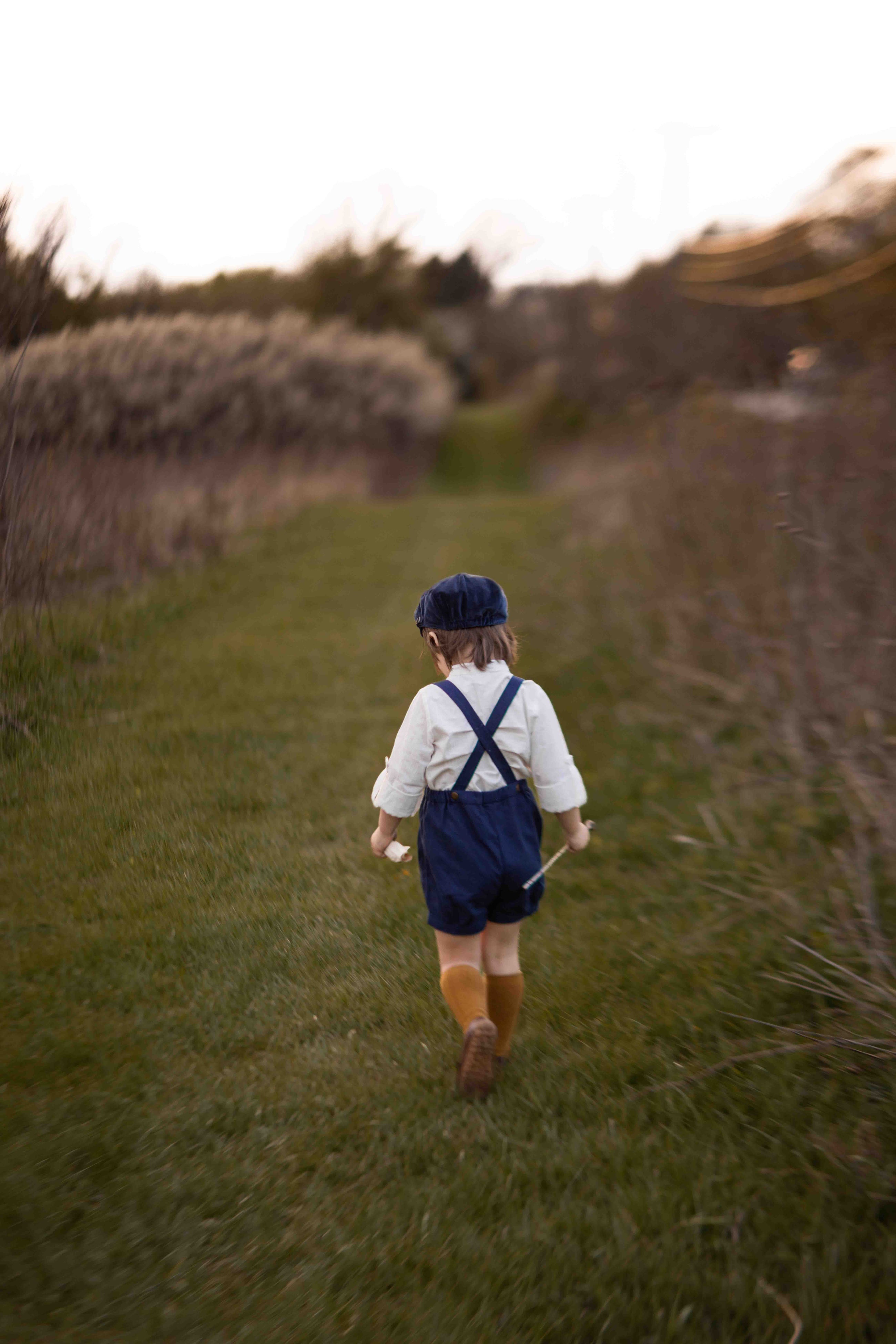 Child walking away in a field wearing a white shirt, blue overalls, and brown boots.