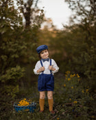 A model boy posing with a smirk, exuding elegance while wearing his navy linen suspender shorts and white Henley shirt. His confident expression and stylish outfit create a charming and sophisticated look.