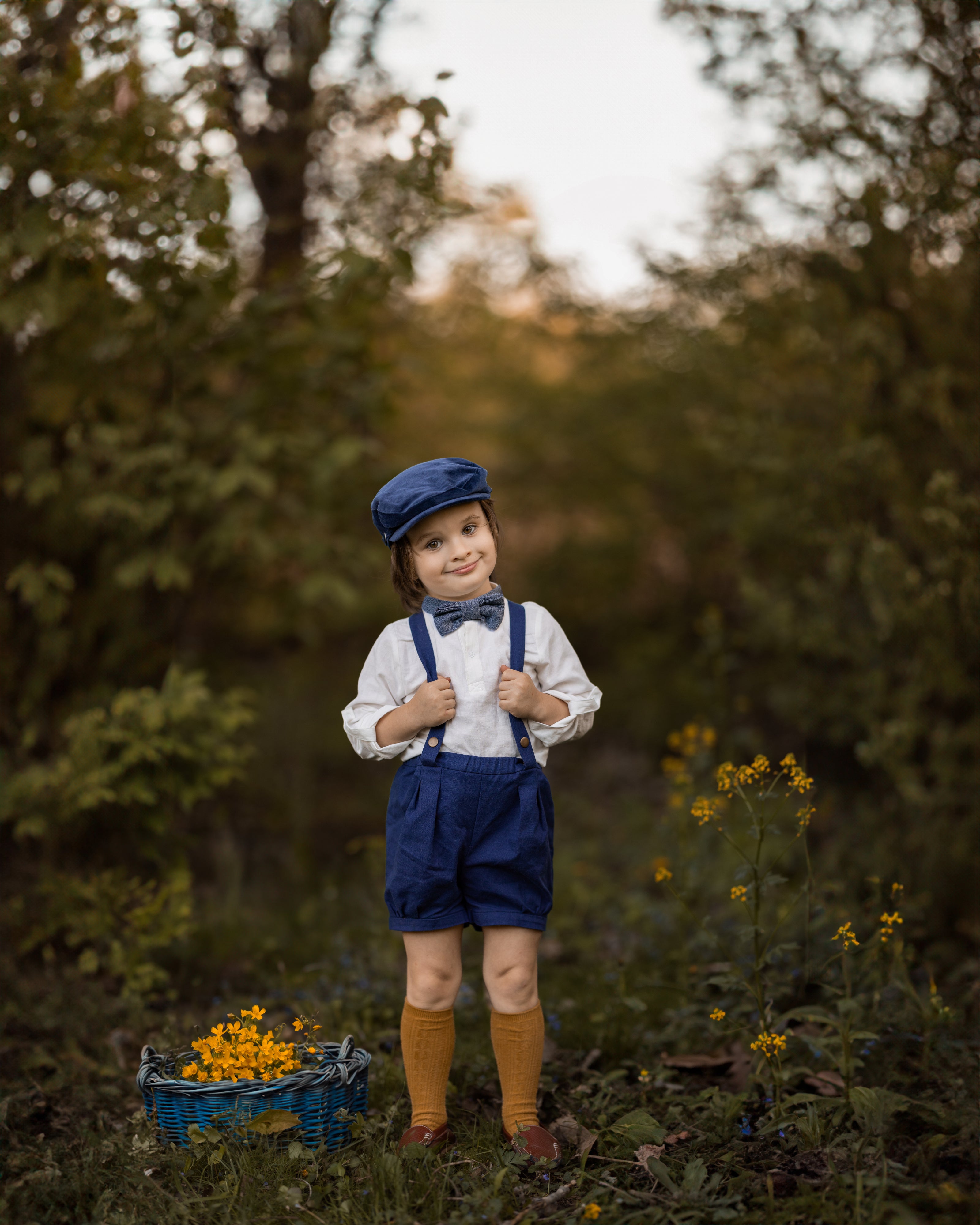A model boy posing with a smirk, exuding elegance while wearing his navy linen suspender shorts and white Henley shirt. His confident expression and stylish outfit create a charming and sophisticated look.