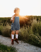 Energetic 3-year-old boy in chambray blue linen short overalls, captured mid-jump, demonstrating the freedom of movement and all-day comfort for active children
