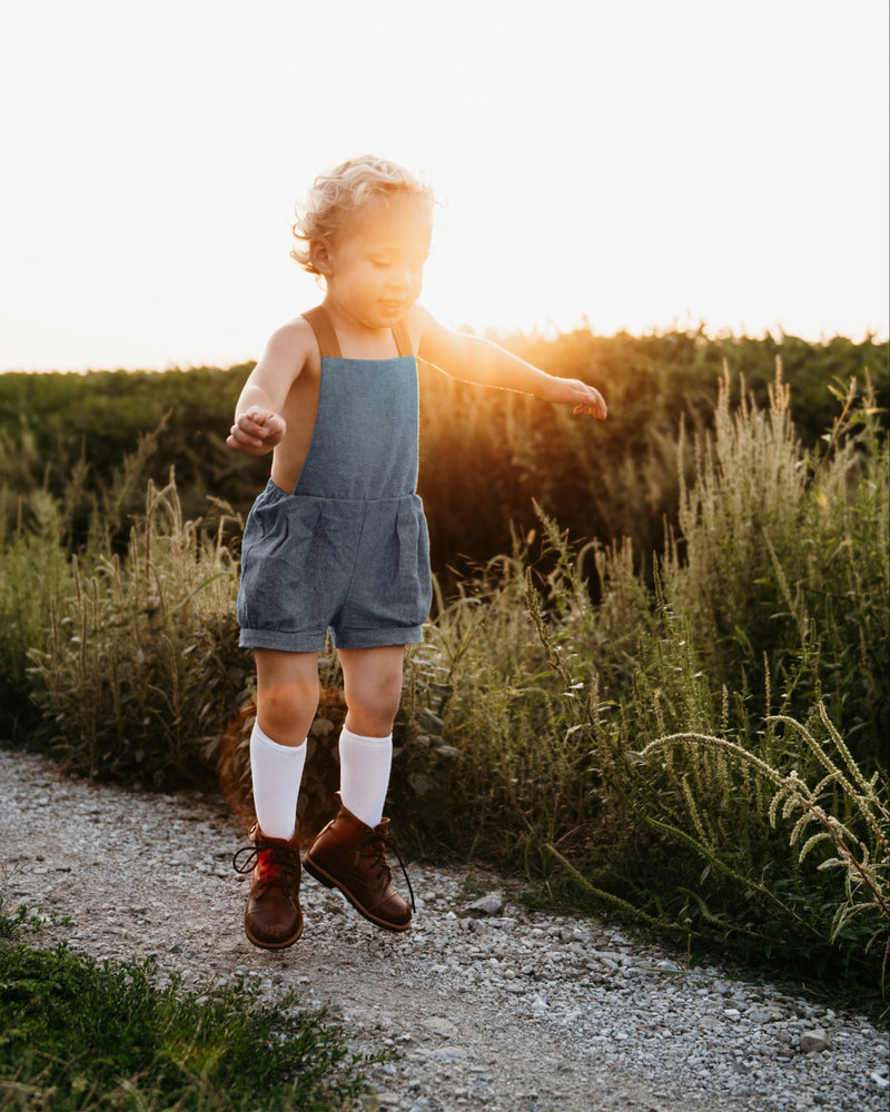Energetic 3-year-old boy in chambray blue linen short overalls, captured mid-jump, demonstrating the freedom of movement and all-day comfort for active children