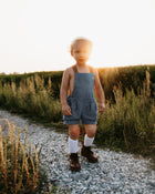 A joyful 3-year-old boy wearing chambray blue linen short overalls, playing in a sunlit field, highlighting the comfort and versatility of natural, sustainable clothing for kids.