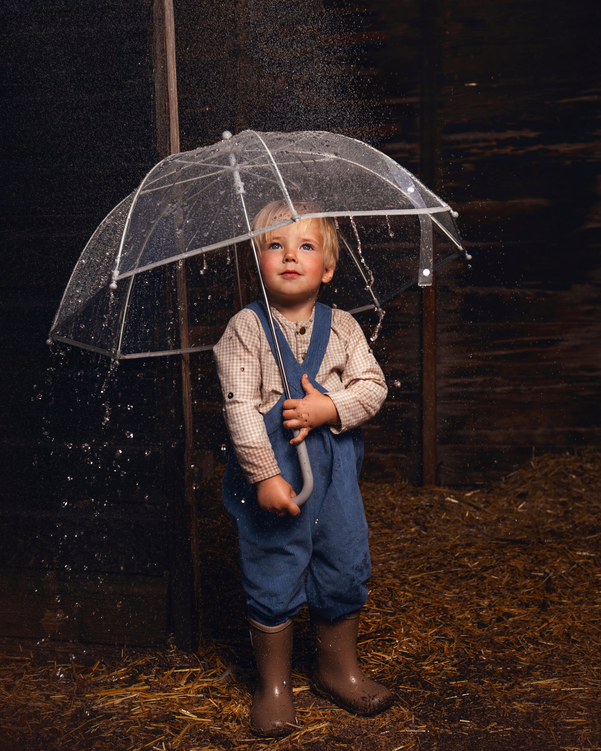 A young child standing in the rain under a clear transparent umbrella, wearing blue overalls, a light patterned shirt, and brown rain boots on a straw-covered floor against a dark wooden background.