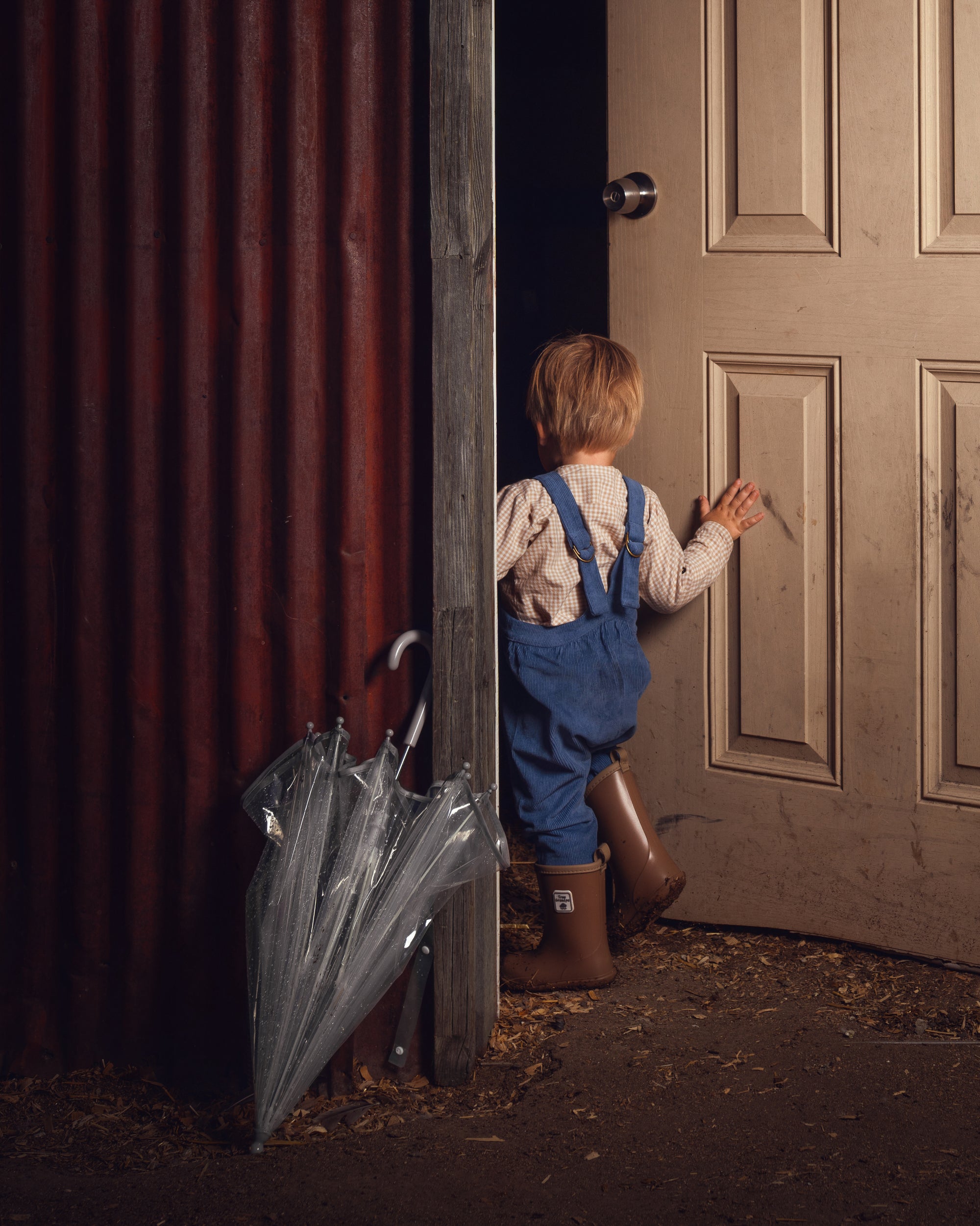 A young child in blue overalls and brown rain boots stands in a doorway, holding a clear plastic umbrella next to a dark red corrugated metal wall.
