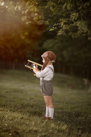 Child in green suspender shorts holding a toy airplane in a natural setting with trees and grass.